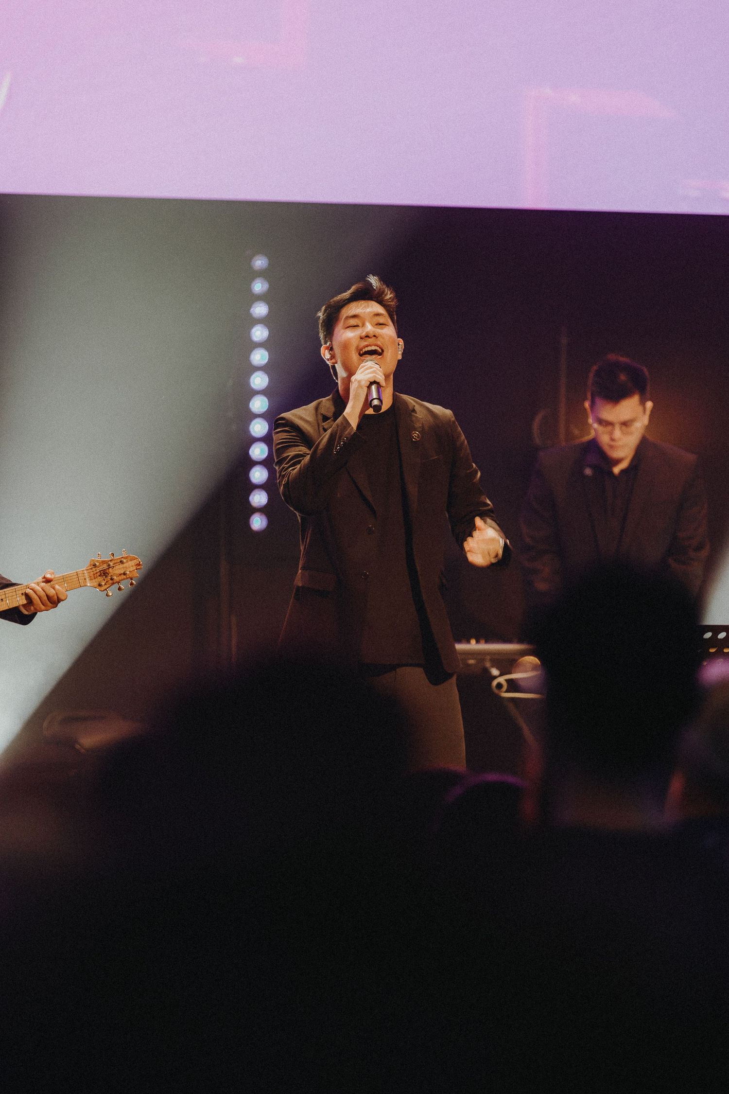 A vocalist performs on stage in a dark suit while backed by musicians during a concert performance.