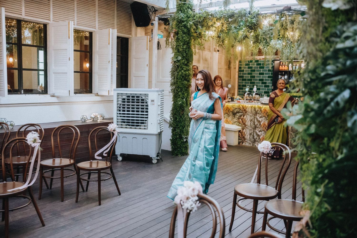 Women in elegant saris walk through a decorated outdoor venue with wooden chairs and lush greenery.