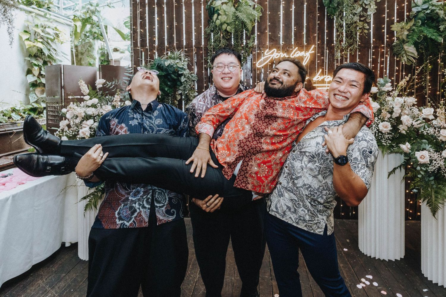 Four men in batik shirts playfully lifting one guest at a festive wedding celebration.