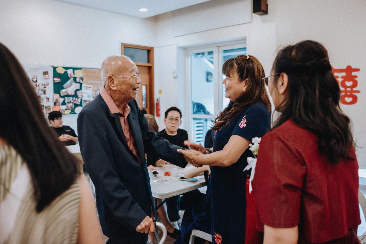 Elderly man greeted warmly by woman in floral dress at a traditional Chinese wedding reception.