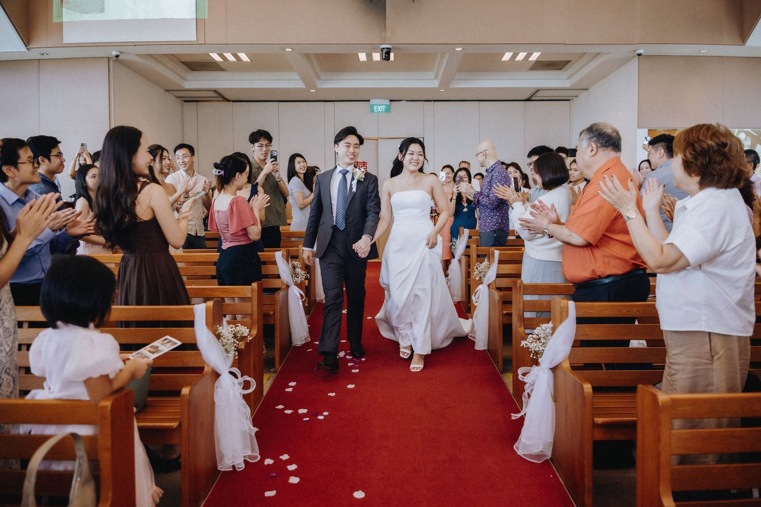 Bride and groom exit church down red petal-lined aisle as guests celebrate their marriage.