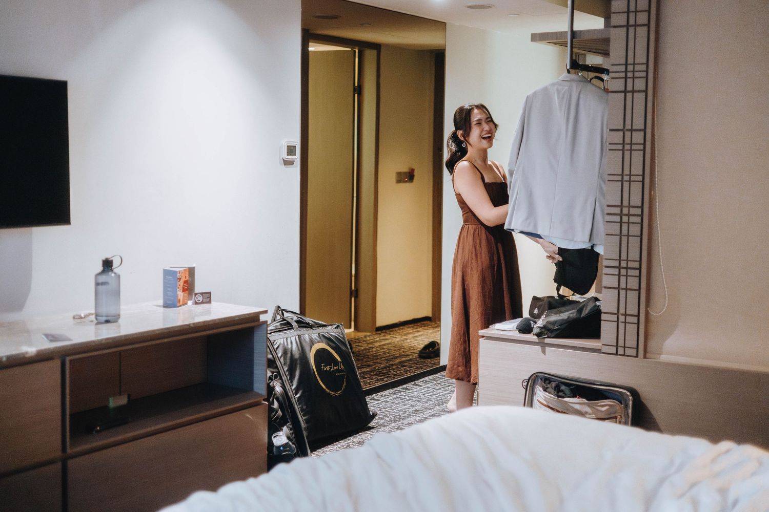 Woman in a rust dress examines hanging wedding attire in a modern hotel room with luggage and TV nearby.