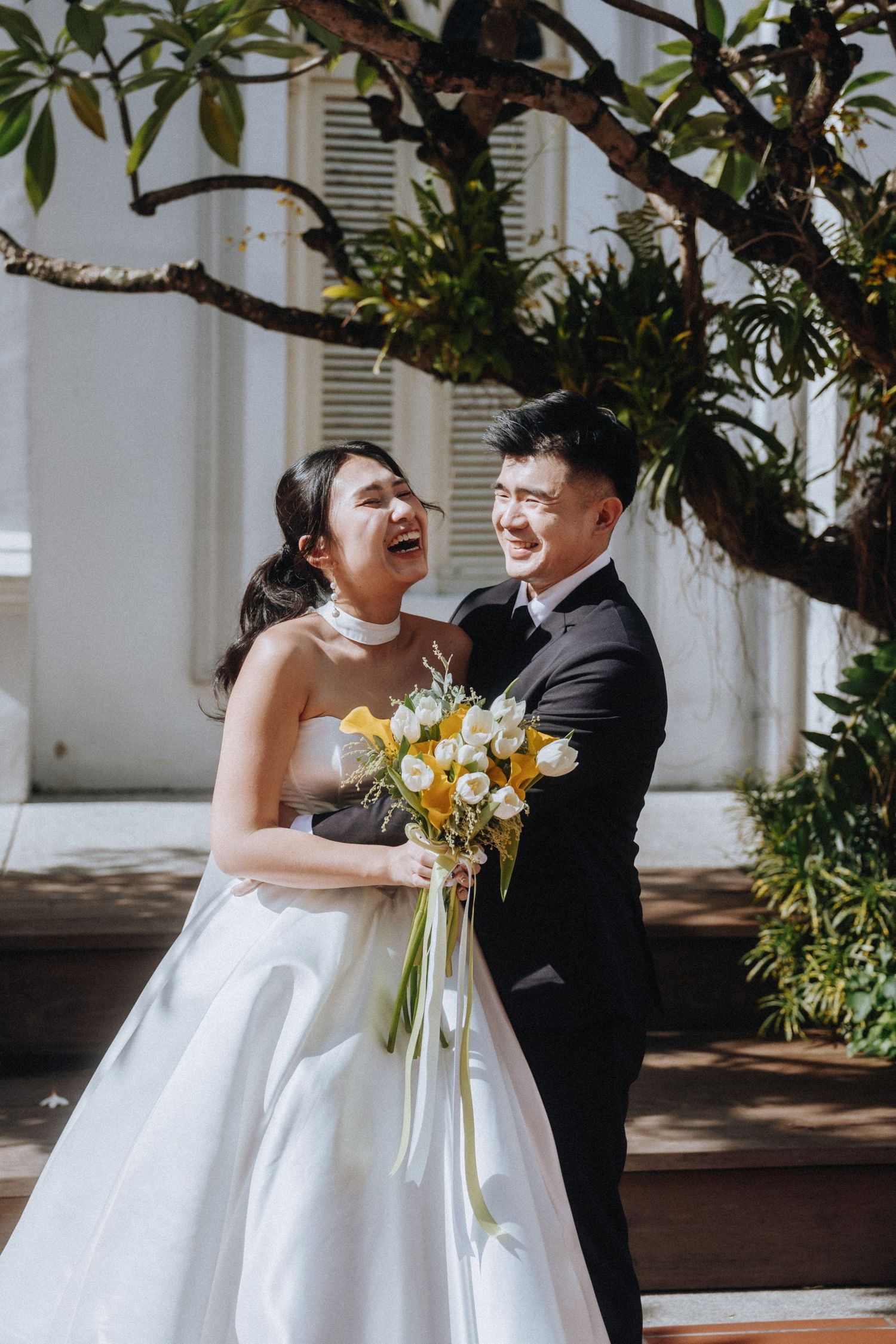 Joyful Asian bride and groom laugh together outdoors holding yellow and white wedding bouquet.