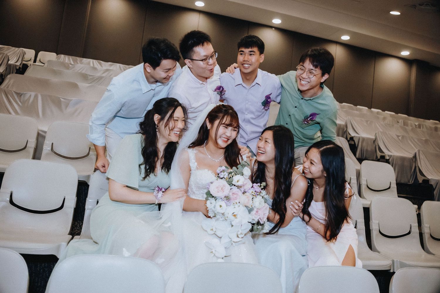 Wedding party members share a joyful moment together in formal attire while seated in white chairs.