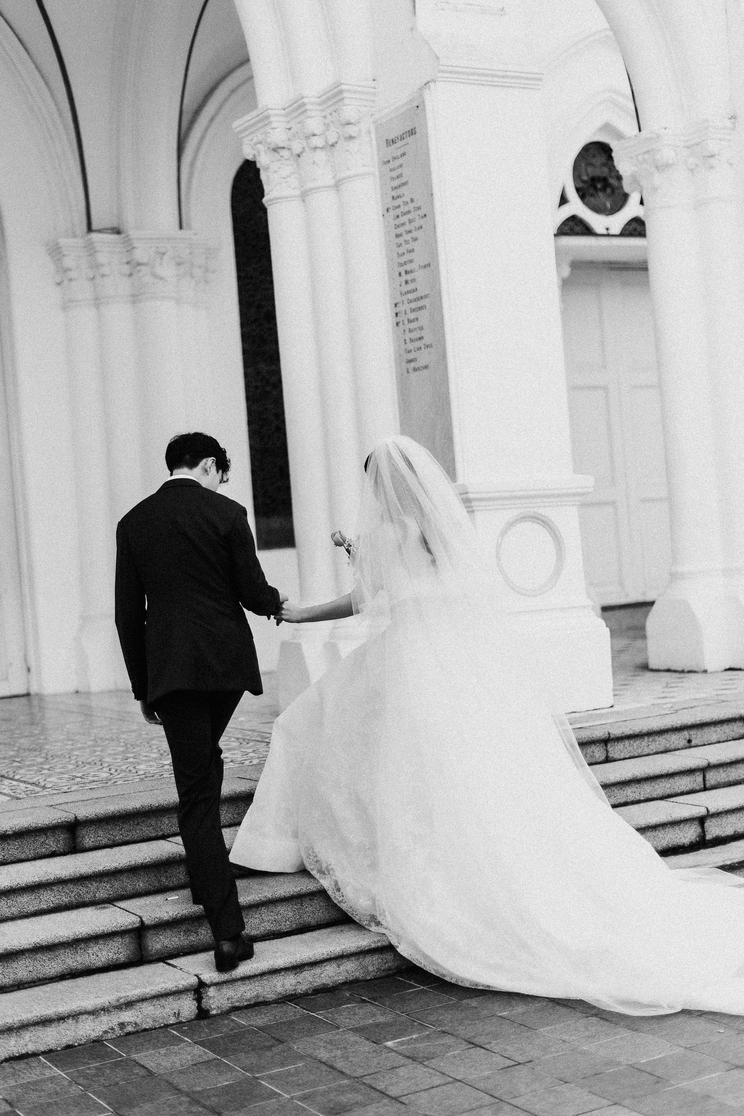 Black and white photo of bride and groom walking up church steps, bride in flowing gown with veil.