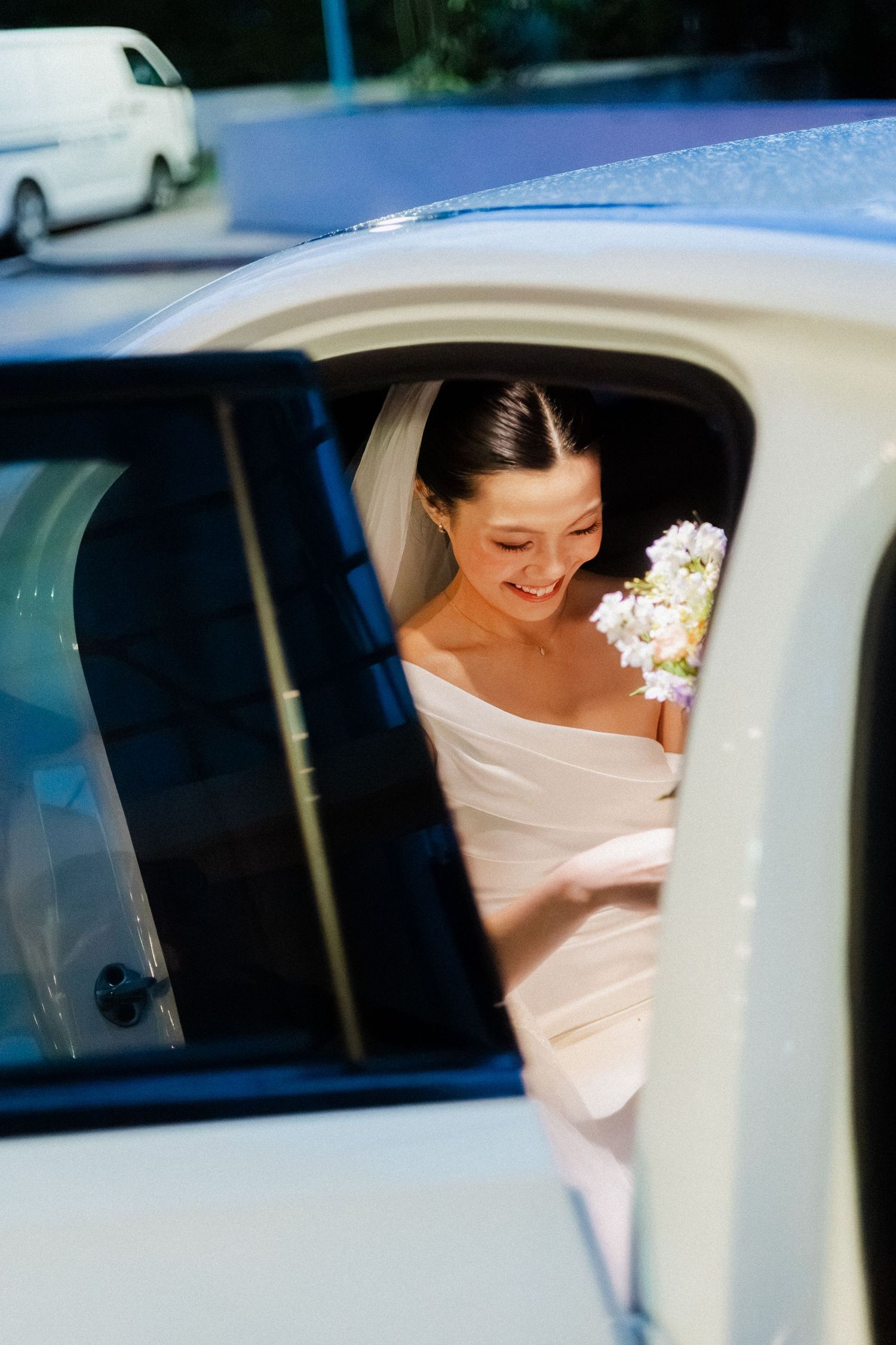 Smiling bride in white gown holding floral bouquet stepping out of a white wedding car.