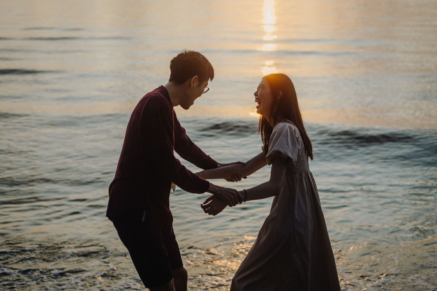 Silhouetted couple holds hands on beach at sunset with ocean waves in background.