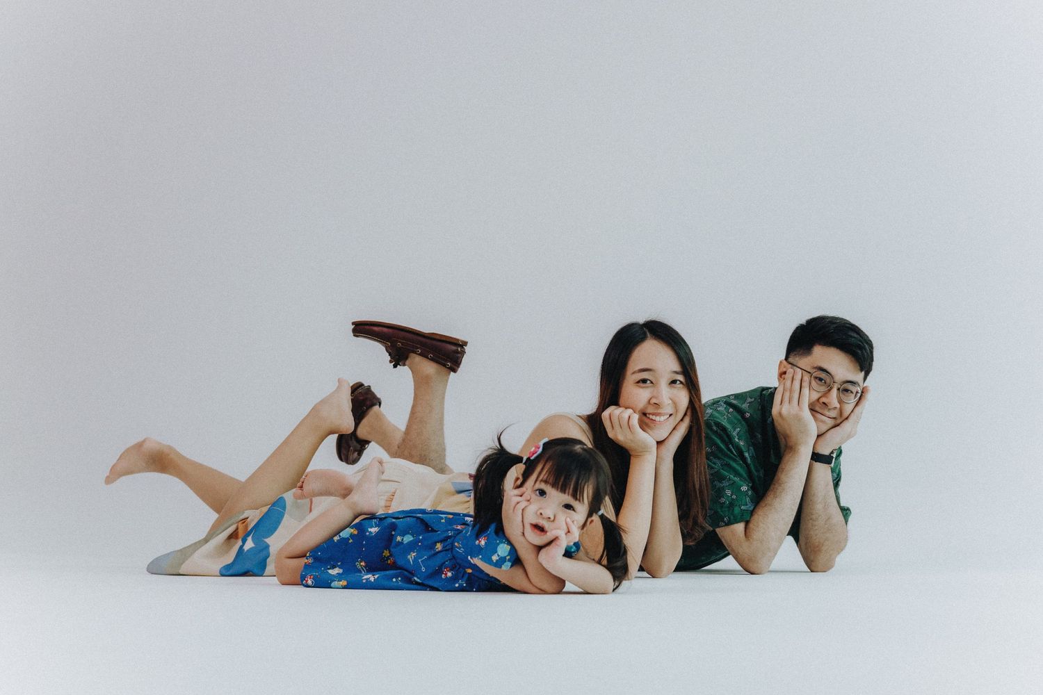 Family lying on white floor in coordinated blue and green outfits for casual portrait session.