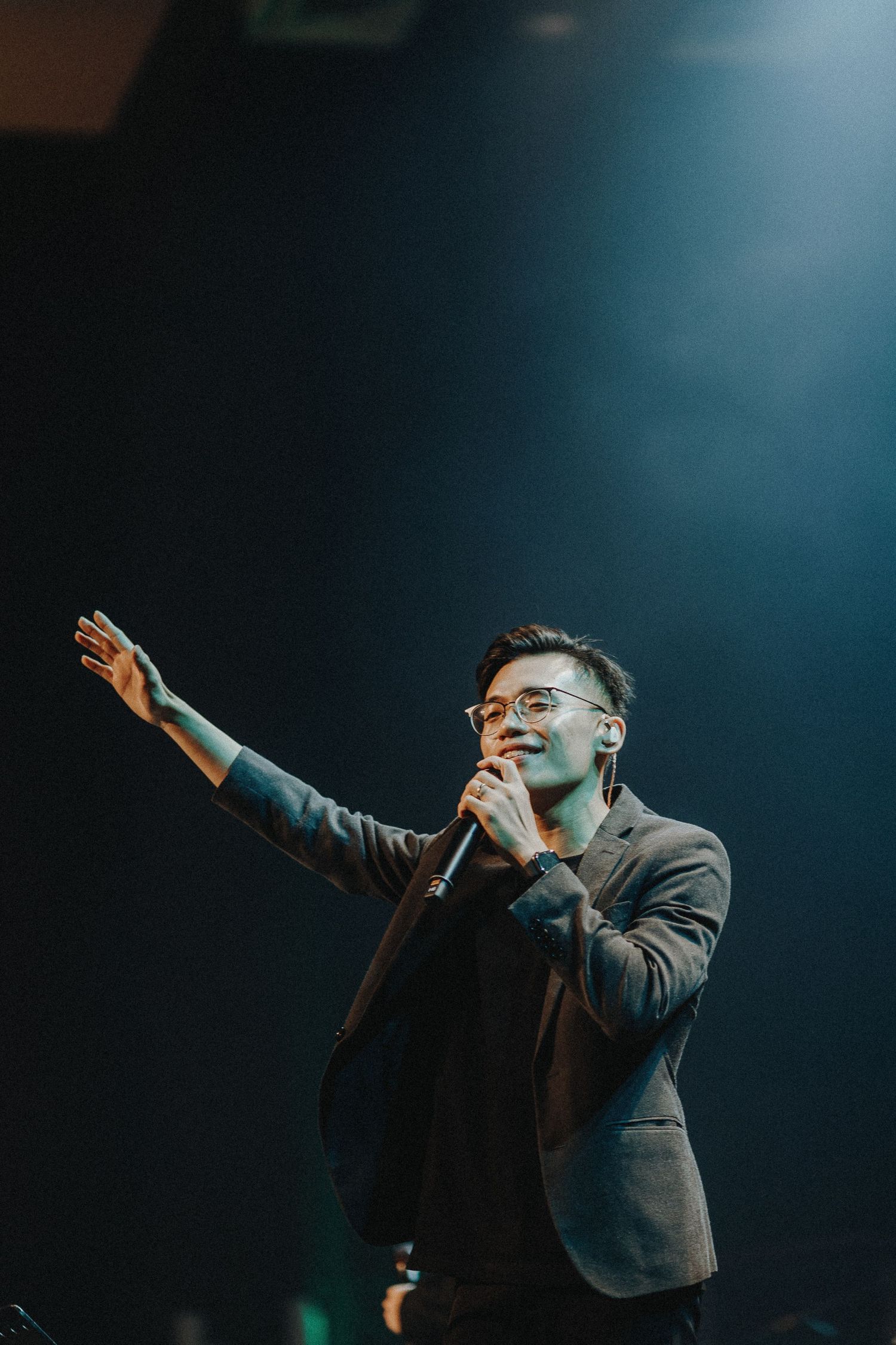 A performer in a dark blazer sings passionately while gesturing on stage under dramatic lighting.