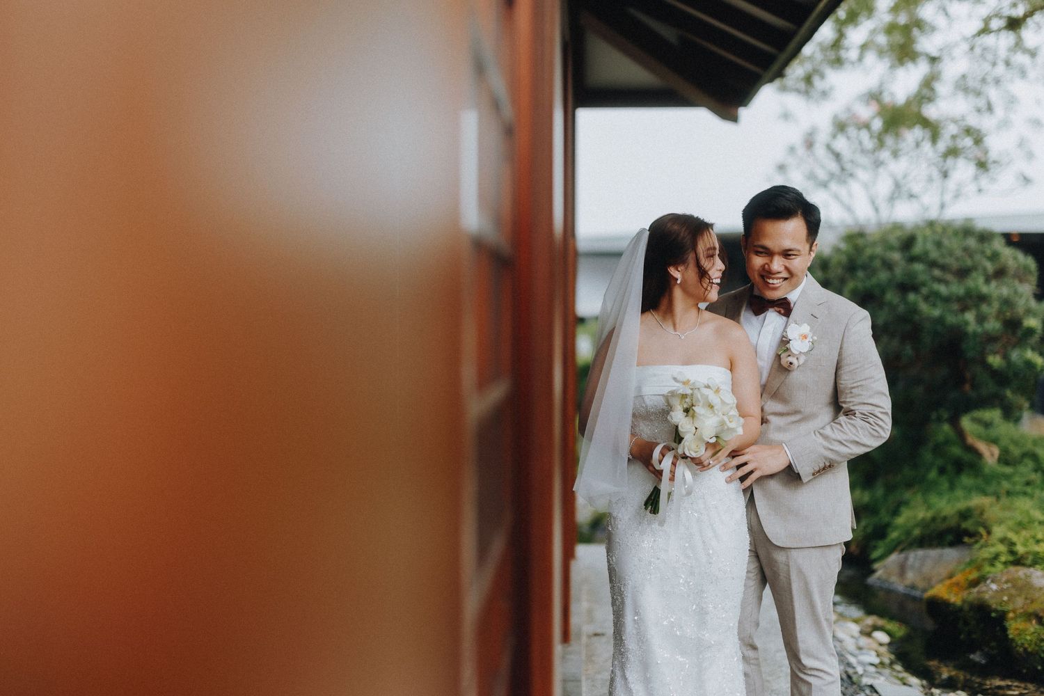 A newlywed couple in light-colored attire poses together against traditional Asian architecture.