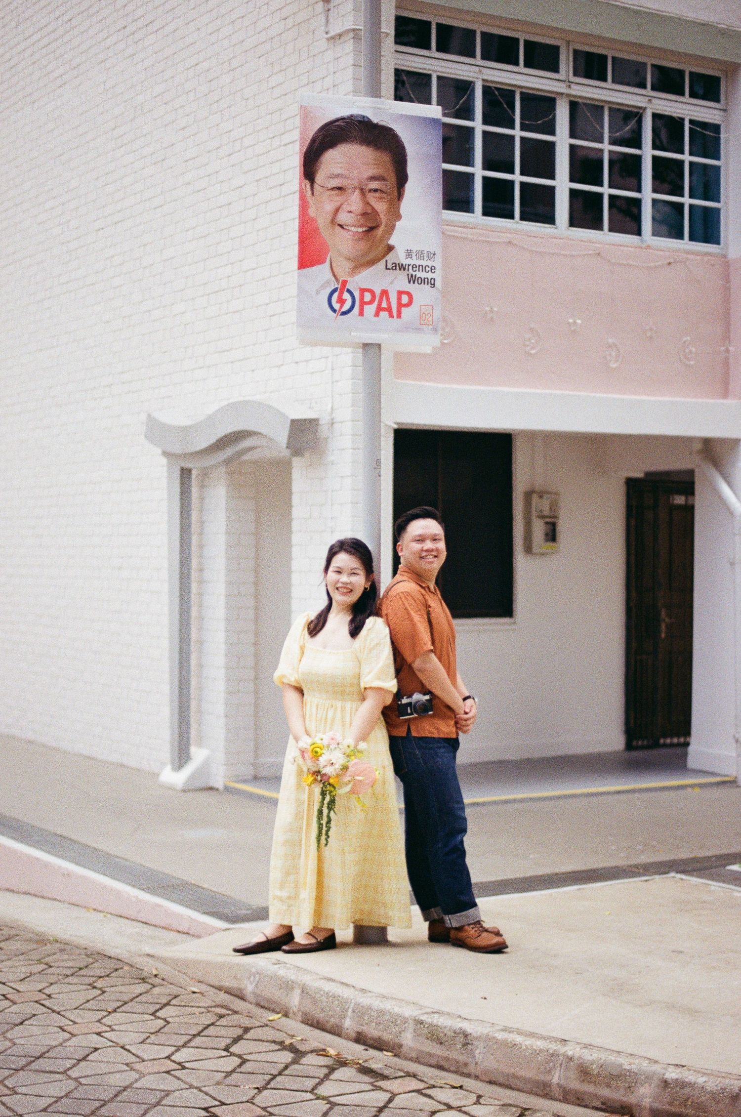 A couple wearing traditional Asian attire poses together in front of a light colored building exterior.