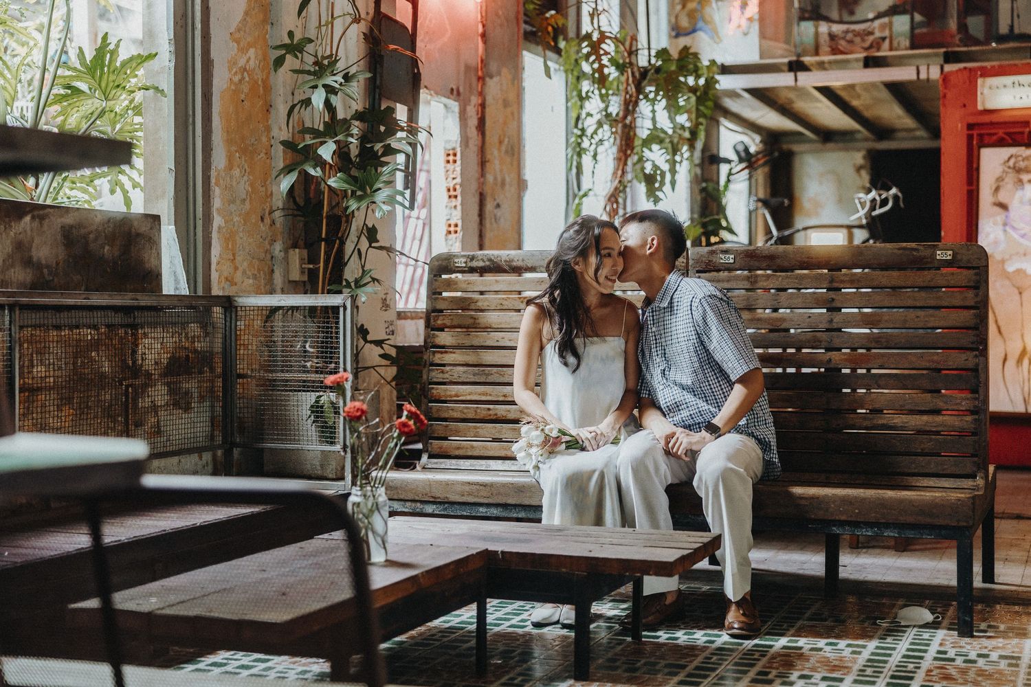 Couple sharing an intimate moment on a bench in a rustic indoor setting with plants and wooden decor.