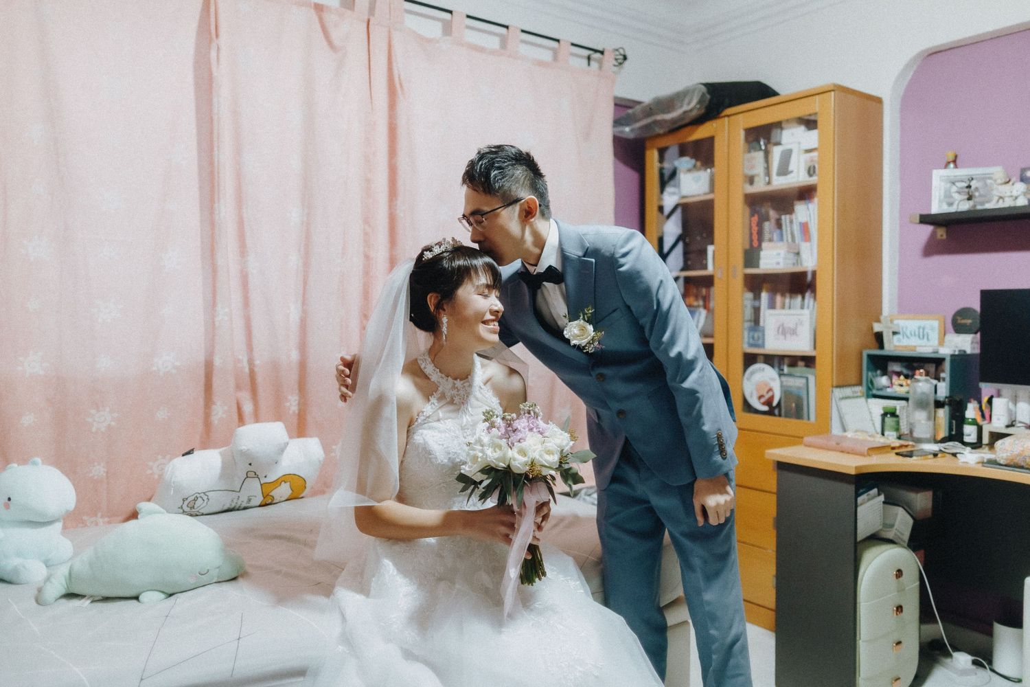 A tender wedding moment in a cozy bedroom with pink walls and bookshelves.