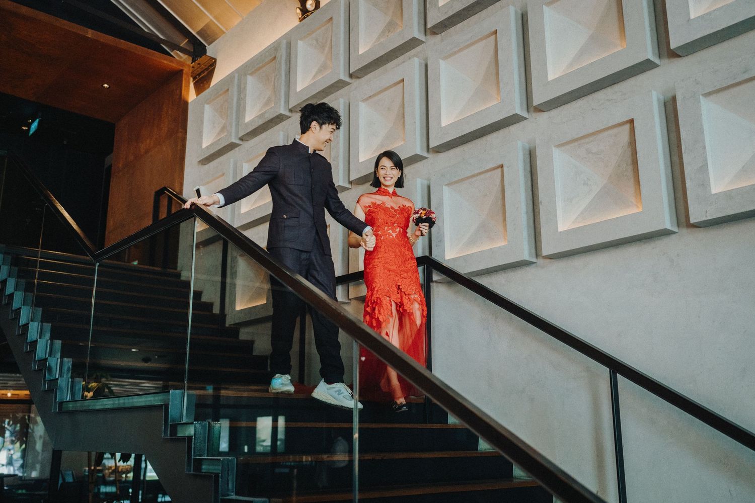 A couple descends an elegant staircase with geometric wall patterns, the woman wearing a striking red dress.