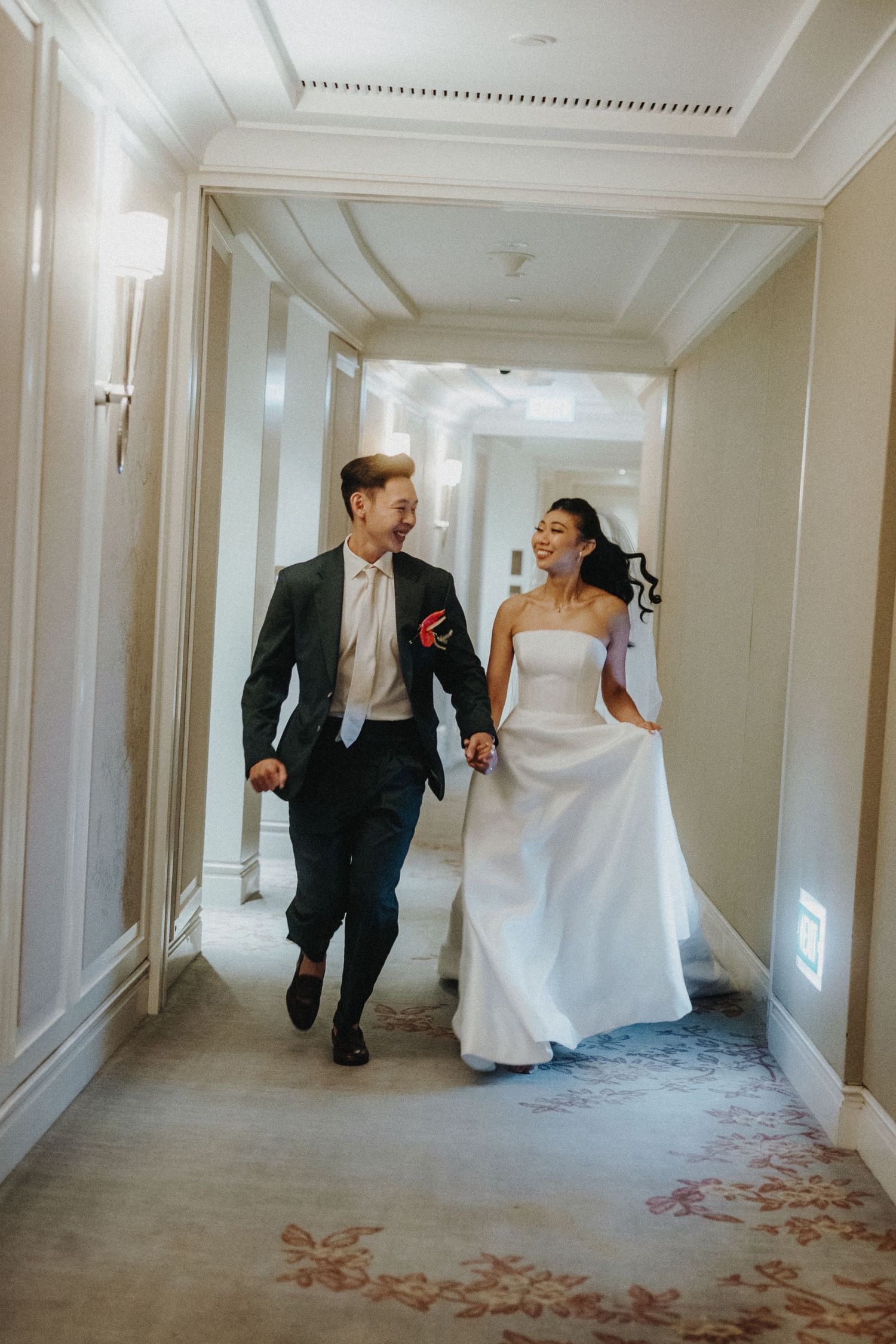 Wedding couple running playfully down an elegant white hotel hallway scattered with rose petals.