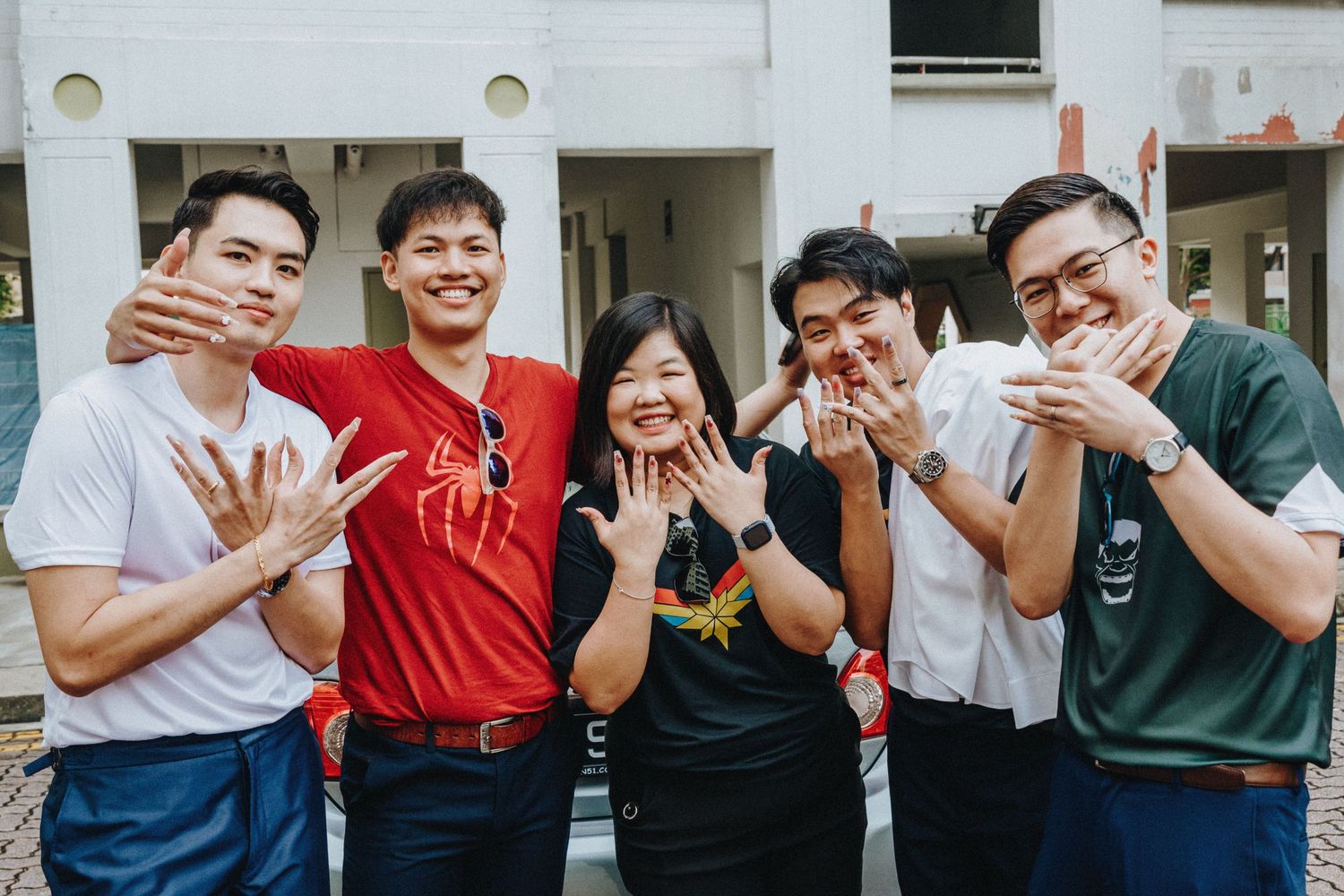 A group of young people posing together outdoors while waving and smiling at the camera.