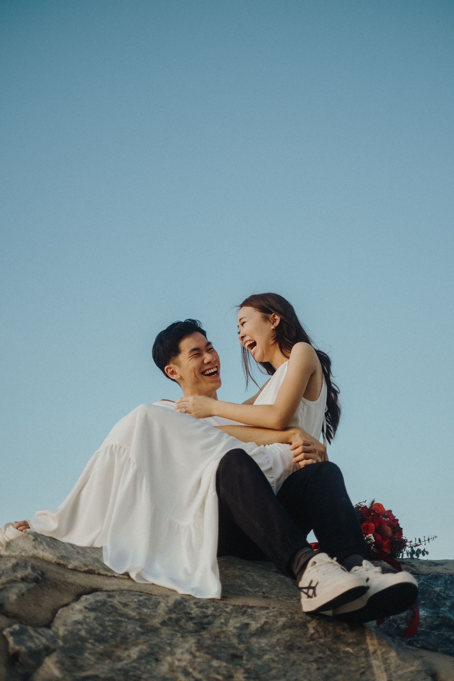 A romantic couple laughs together while sitting on rocky terrain against a blue sky.