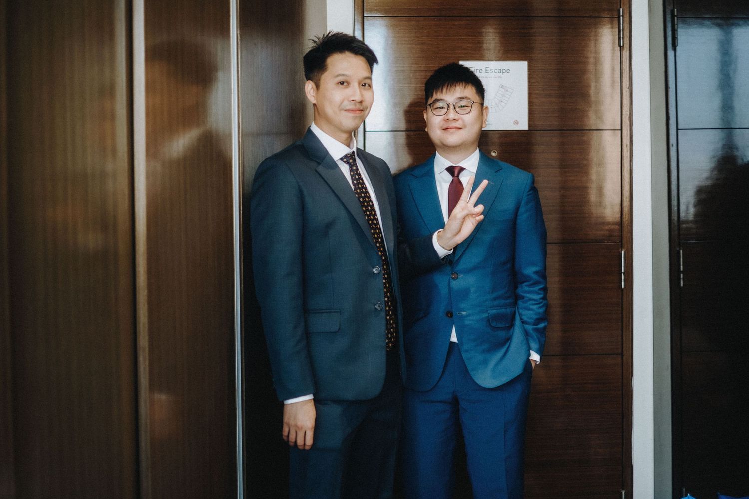 Two men in suits posing for a photo, one making a peace sign, standing near a wooden door.