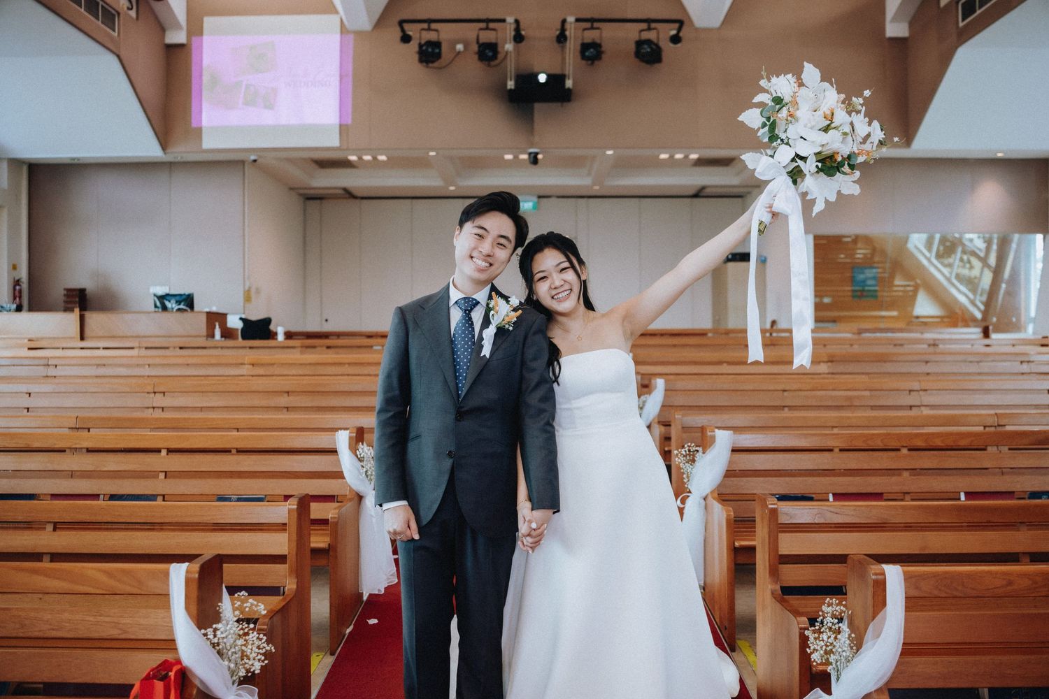 Newlywed Asian couple posing in church aisle, bride in white strapless gown holding bouquet, groom in gray suit.