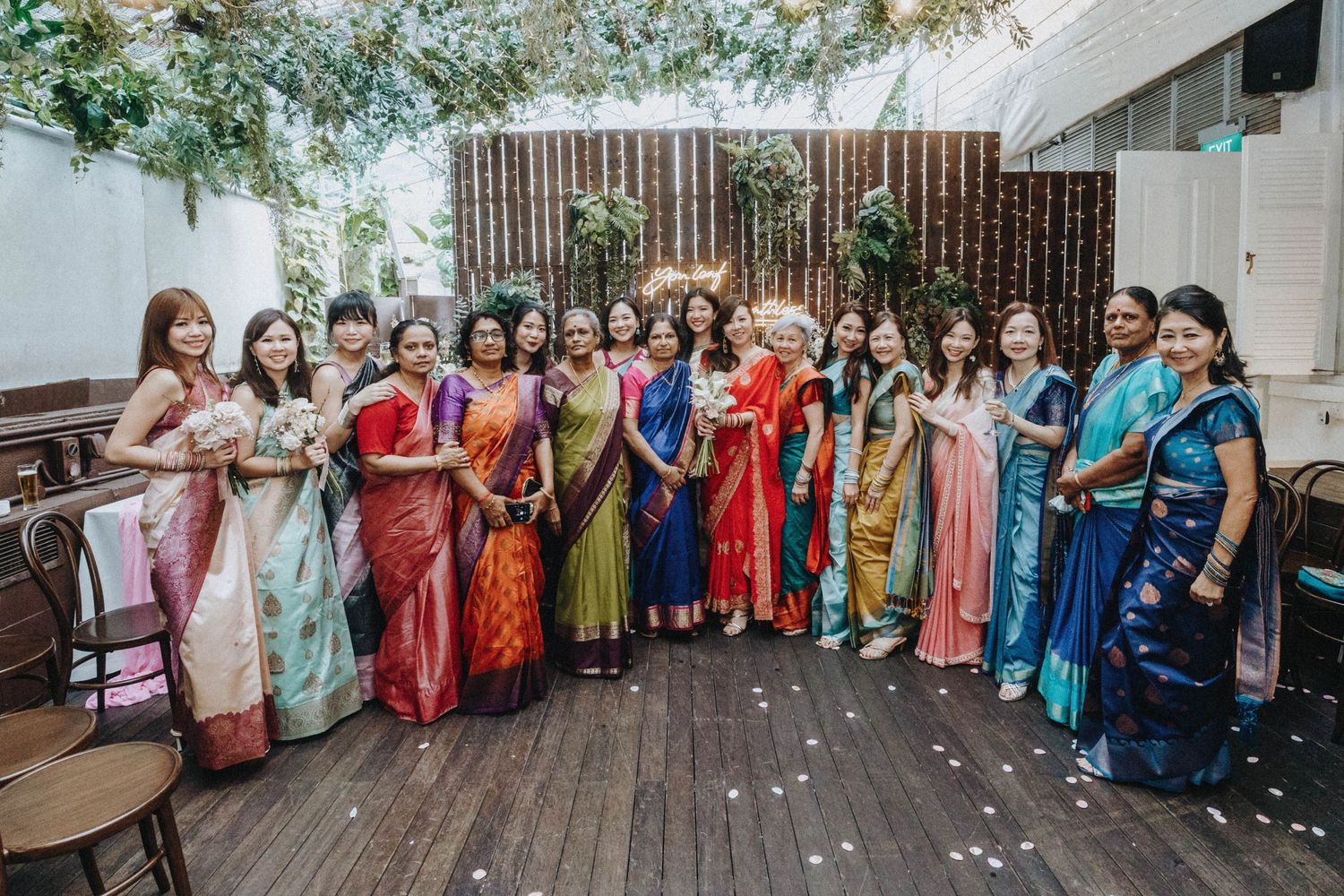 Group of women in vibrant colorful sarees pose together at an elegant garden wedding celebration.