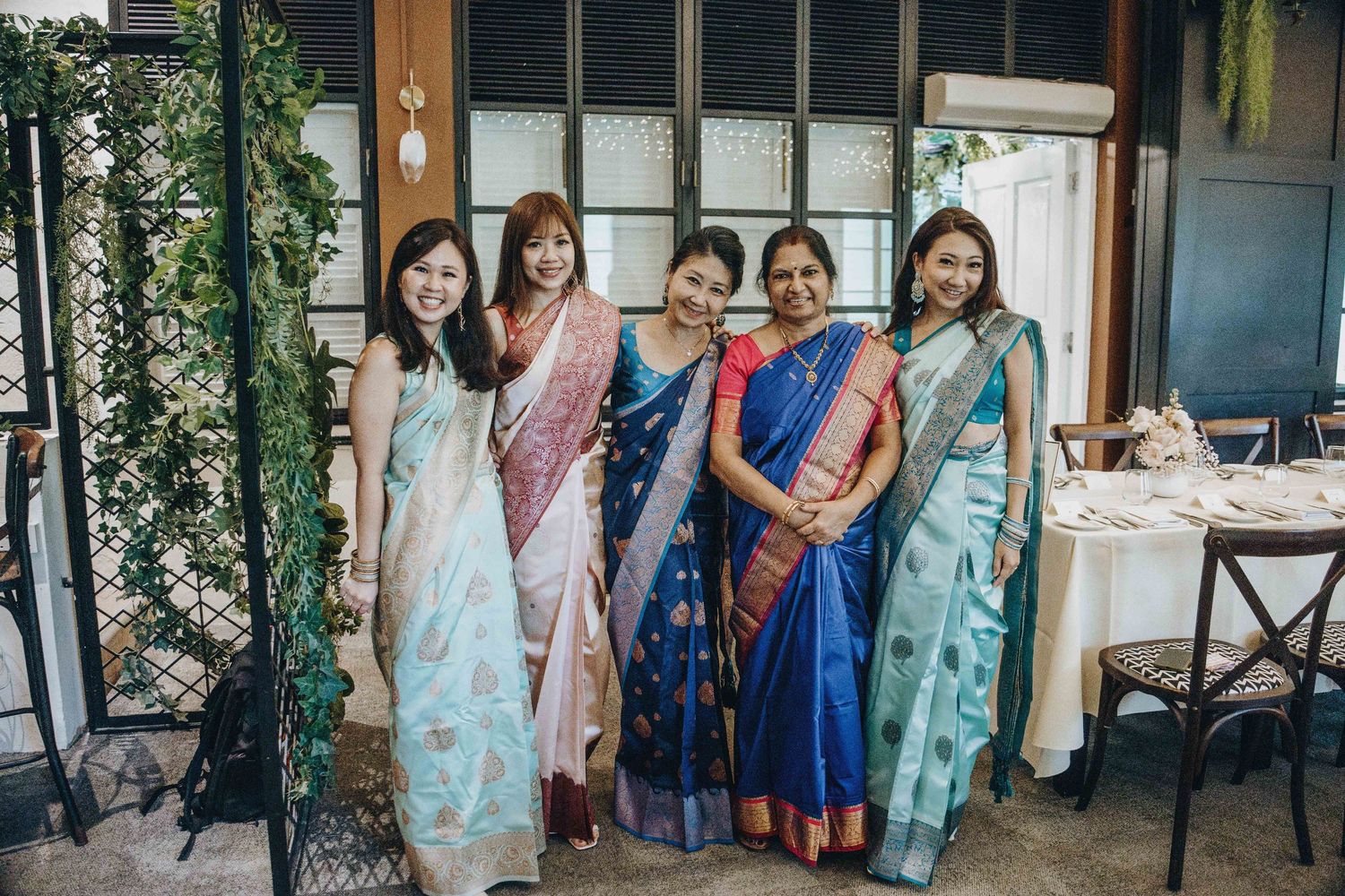 Five women posing together in colorful traditional Indian sarees at an elegant restaurant setting.