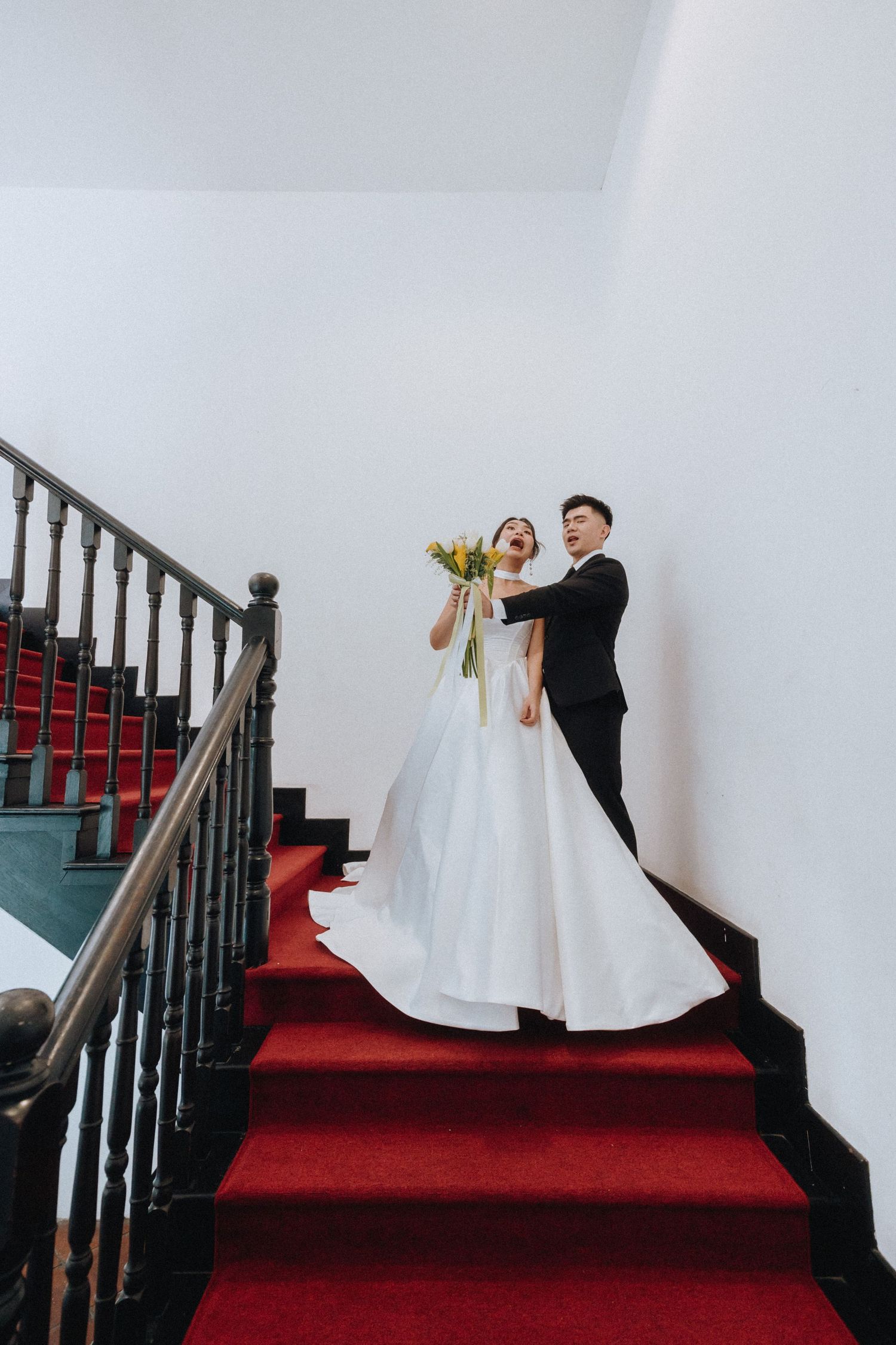 Bride and groom pose on red-carpeted stairs, bride holding yellow bouquet in elegant white gown and groom in black suit.