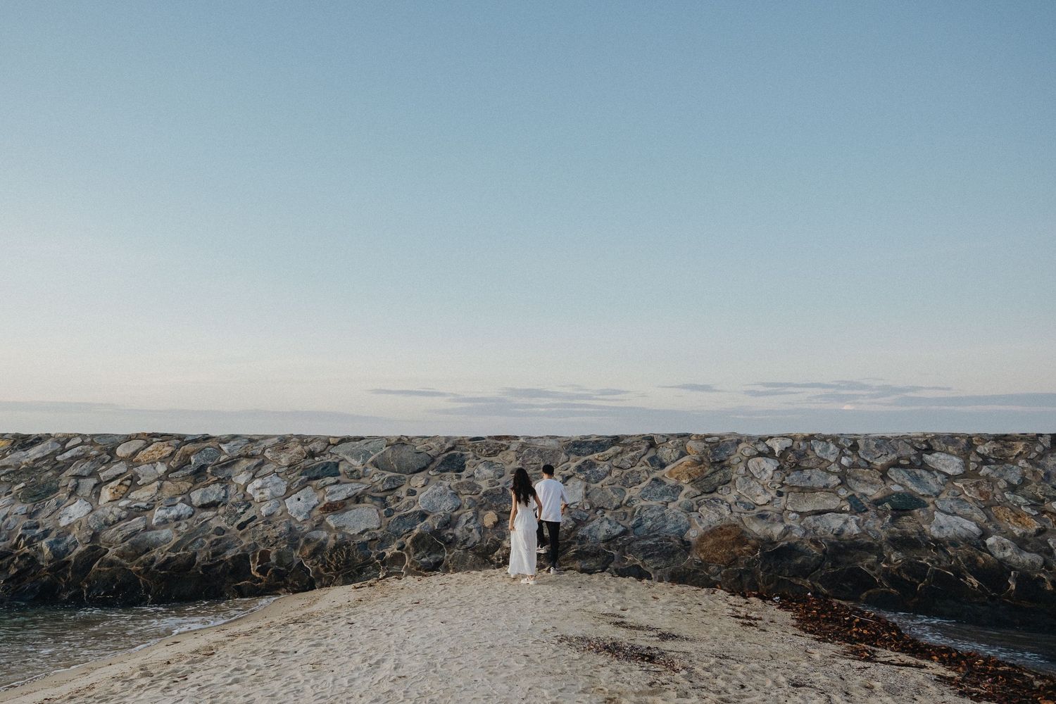 Couple standing on stone jetty by ocean during sunset.