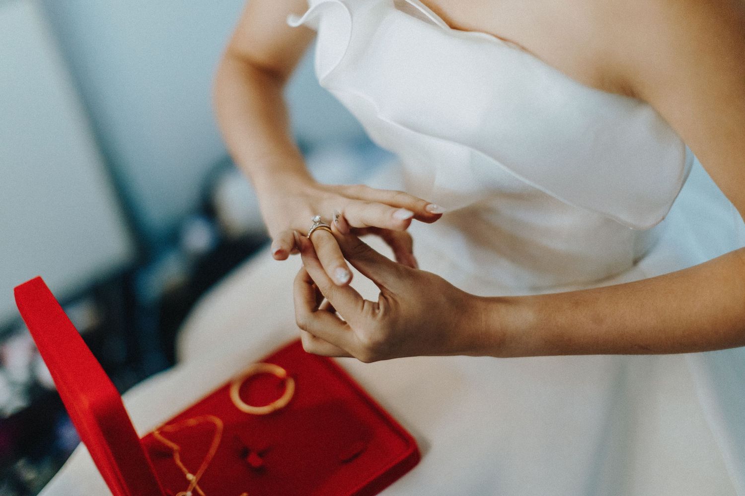 Someone in a white flowing dress holds rings while sitting near a red jewelry box.