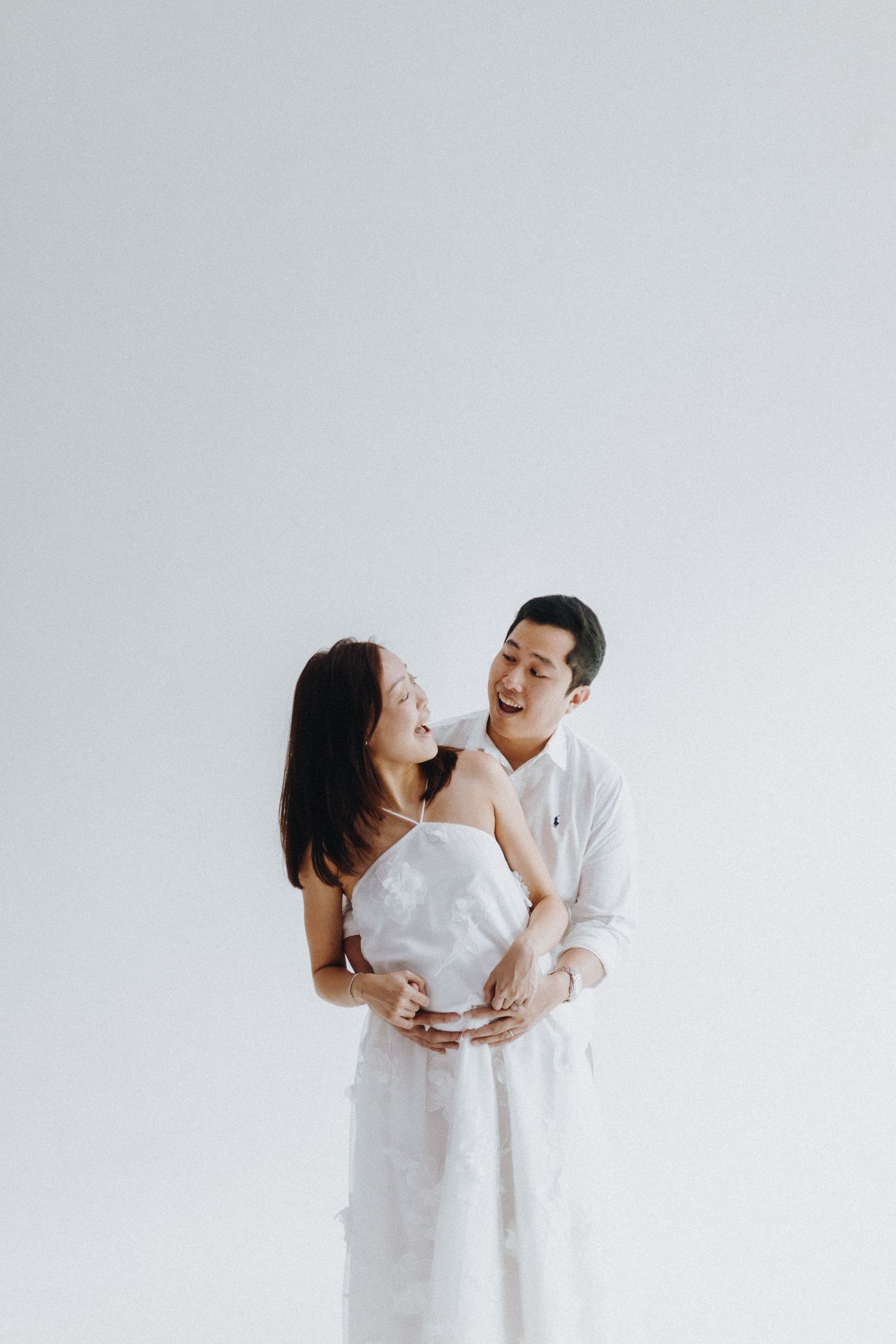 Couple in white outfits sharing a joyful moment against a light background during a studio photoshoot.