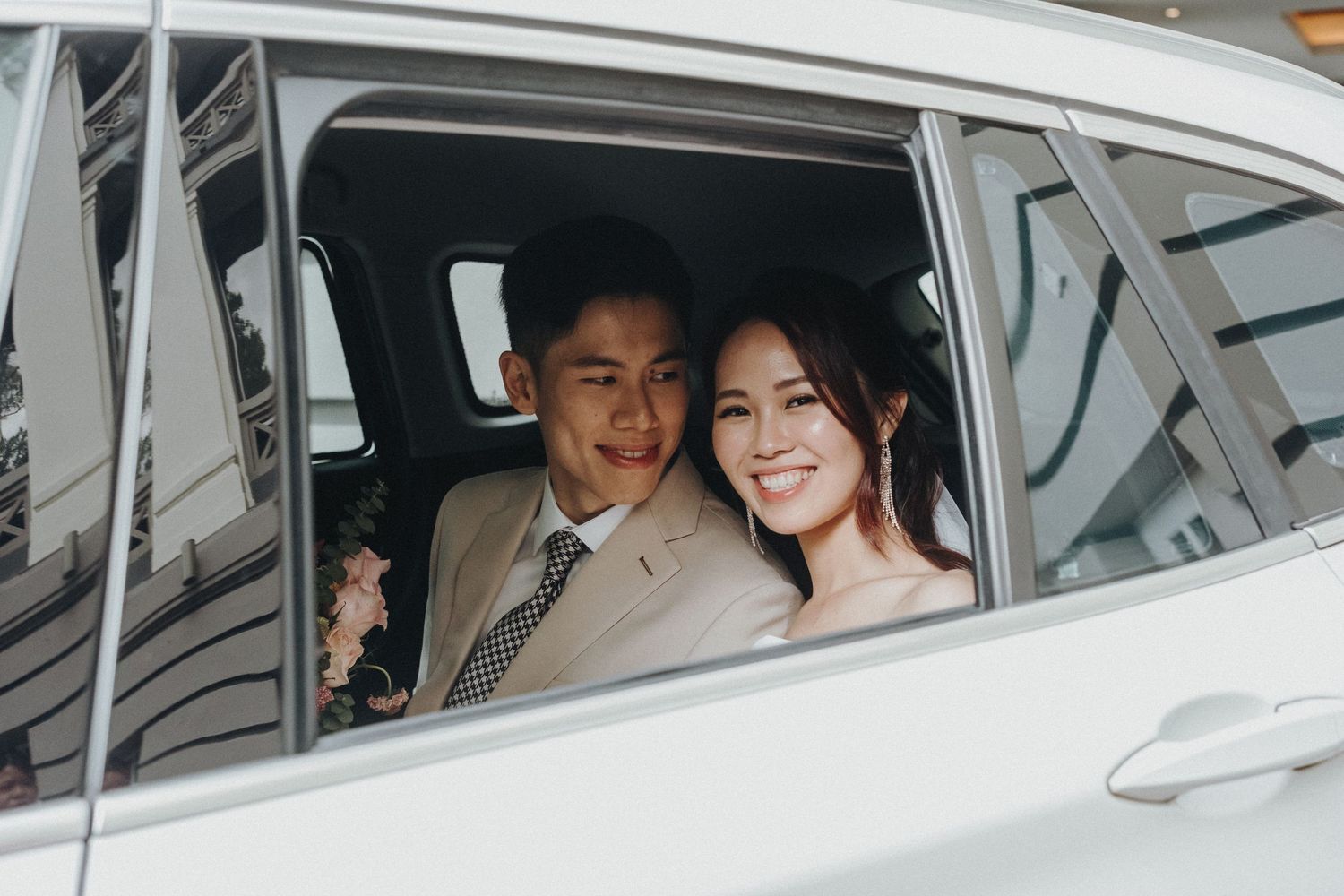A couple sits together in the backseat of a car, smiling through the window on their special day.