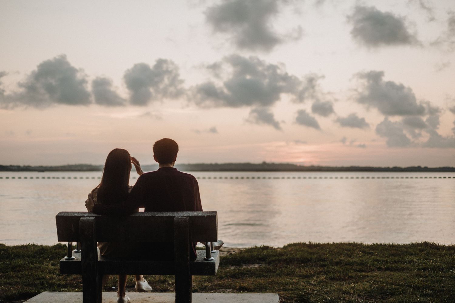 A silhouetted couple sits on a bench overlooking a serene lake at sunset with clouds reflecting on the calm water.