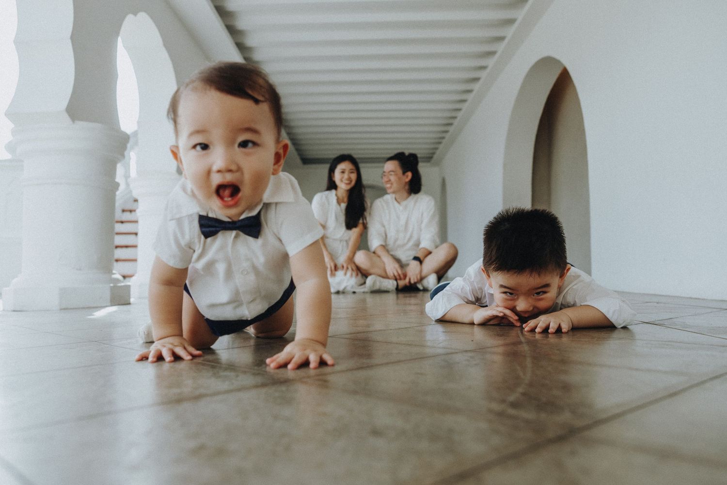 A family plays together in a bright white corridor with arched columns, creating a playful moment on the floor.