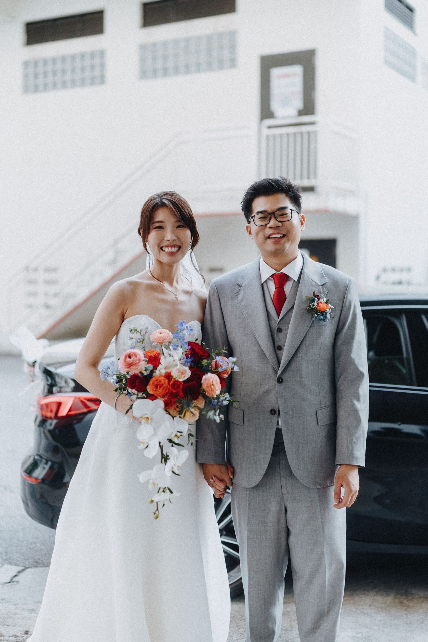 A joyful couple in wedding attire poses together with a vibrant coral and pink bouquet.
