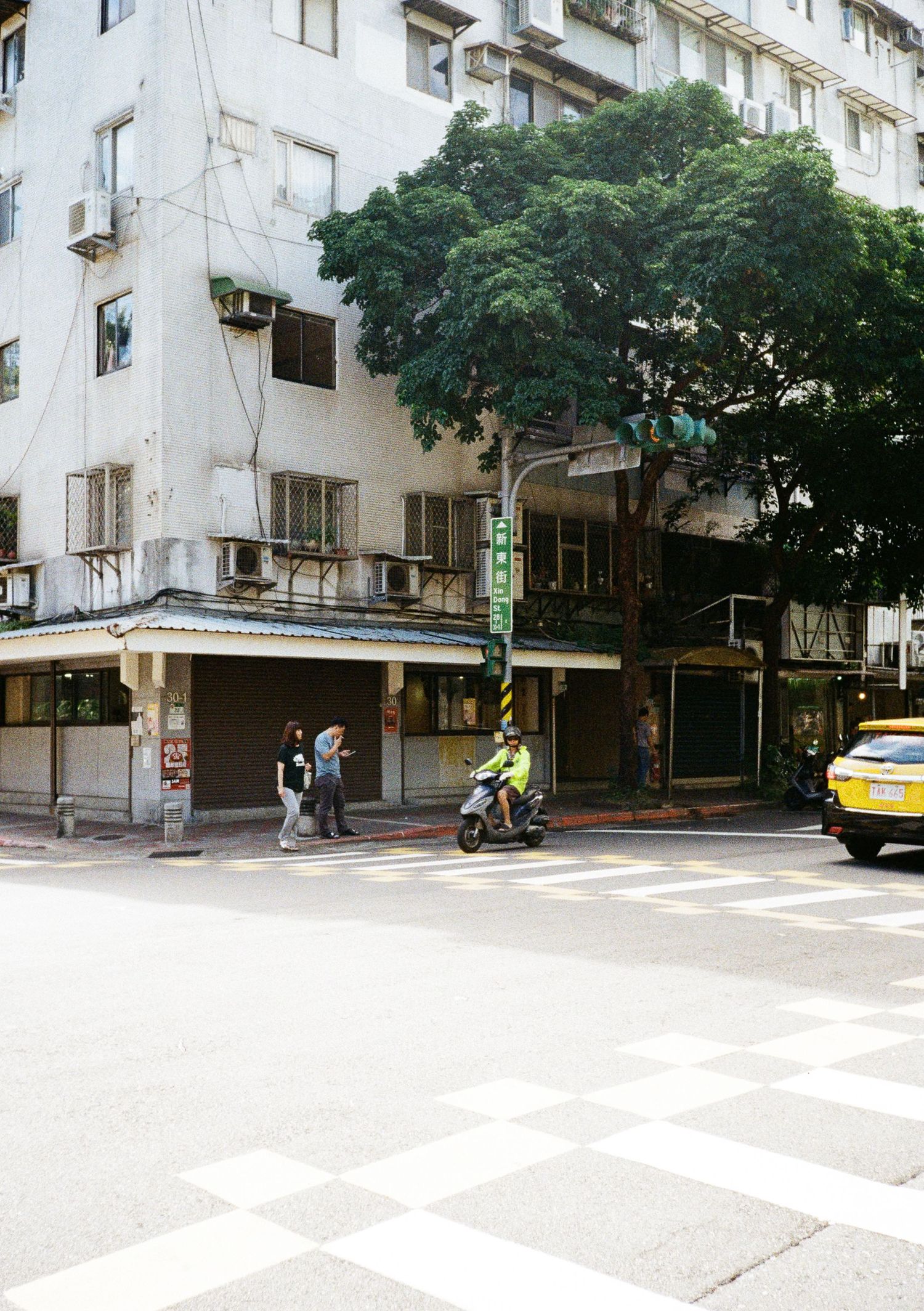 Urban street corner with beige apartment buildings, trees, and a yellow taxi cab visible in front of a crosswalk.