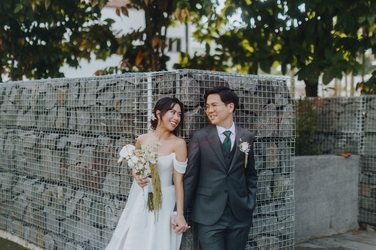 A bride and groom pose together against a modern stone wall backdrop during their wedding photoshoot.