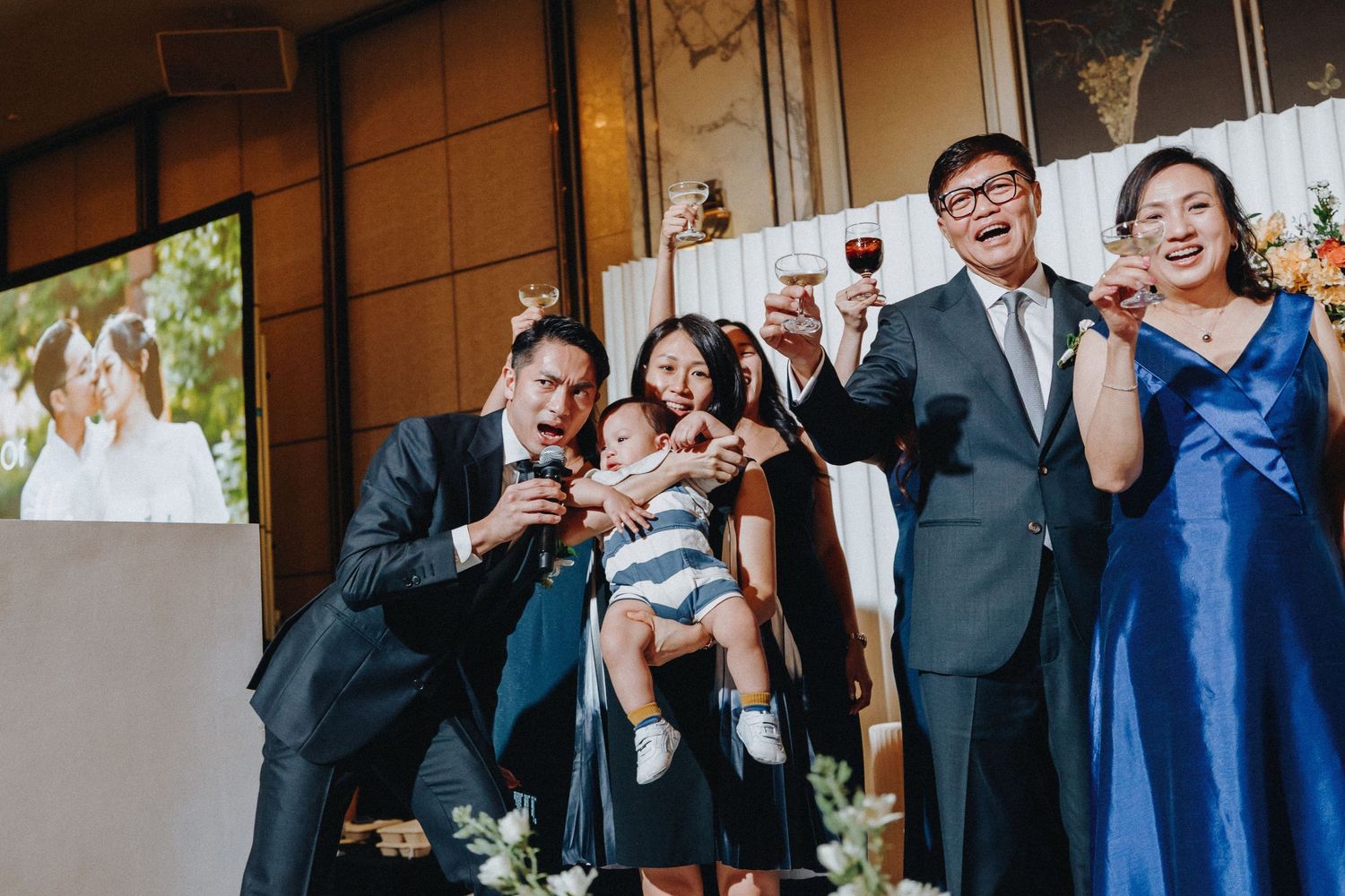 Wedding reception celebration with guests toasting and projection screen in background.