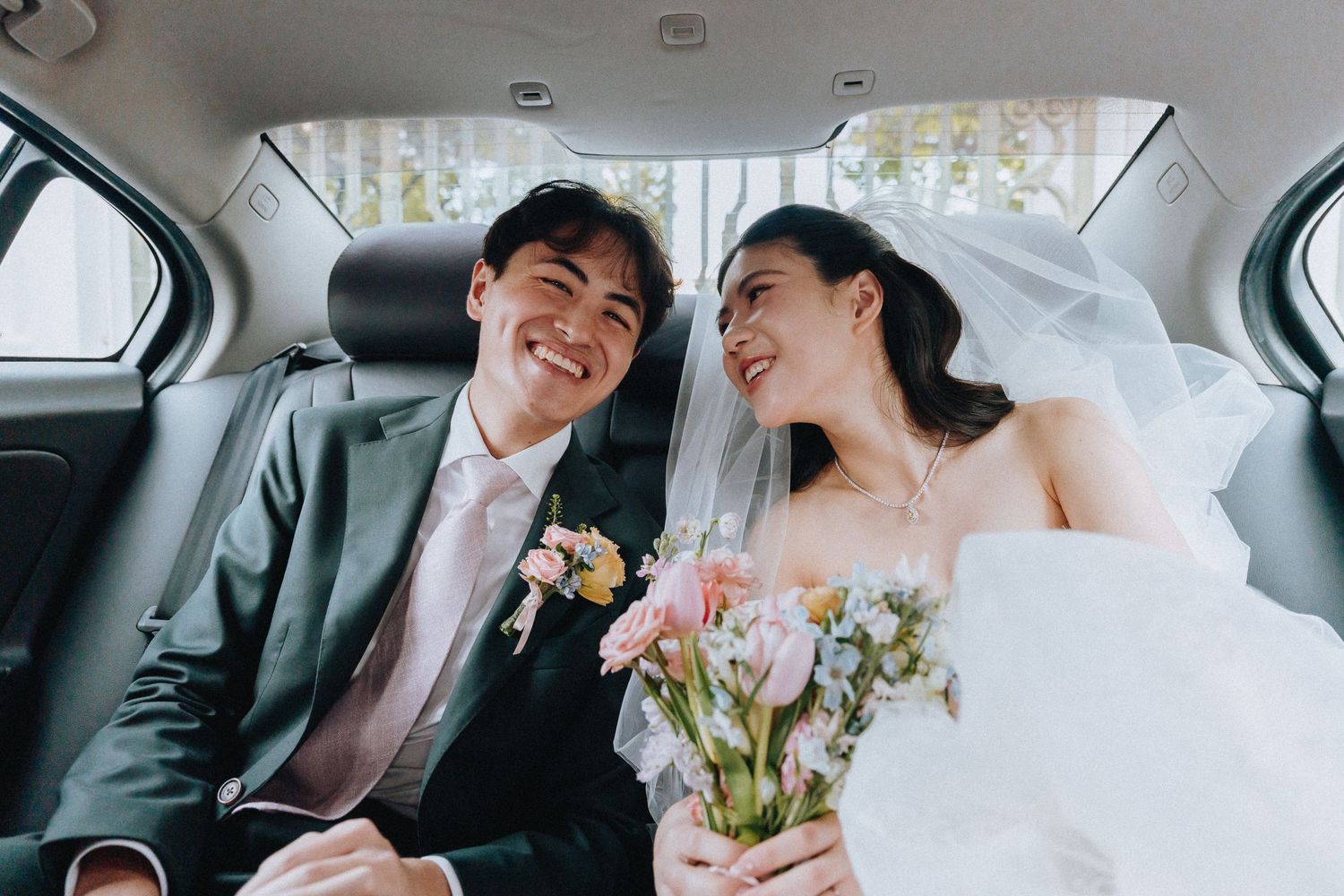 Happy newlywed couple smiling in car backseat, bride in white gown holding bouquet, groom in gray suit.