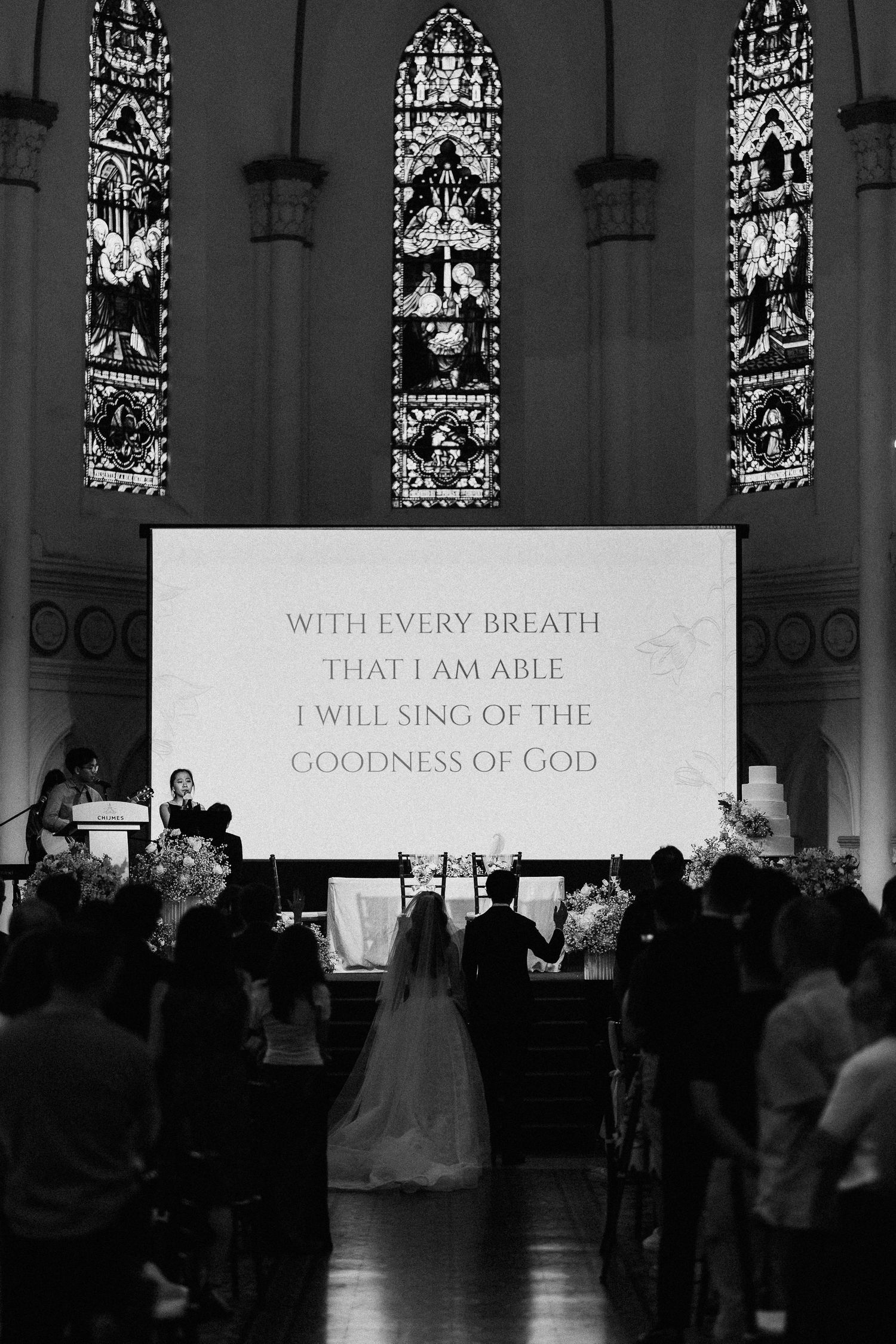 Black and white wedding ceremony in a church with stained glass windows and a screen displaying lyrics about the goodness of God.