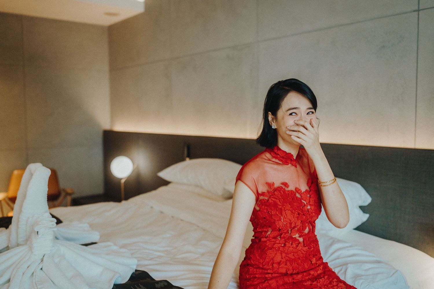 Woman in elegant red lace dress relaxes on white hotel bed linens.