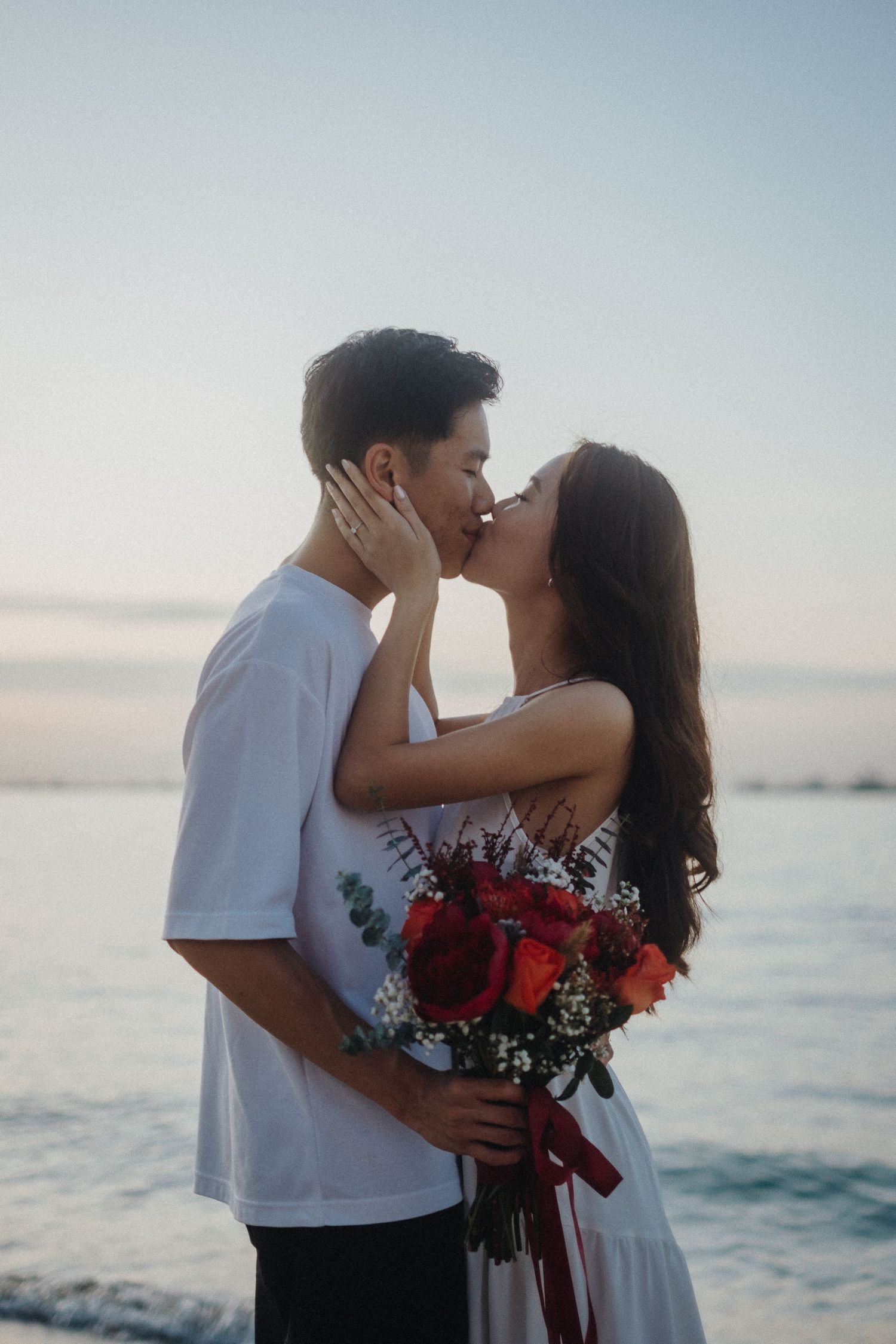 A romantic couple shares a passionate kissing moment at sunset by the ocean with a vibrant red floral bouquet.