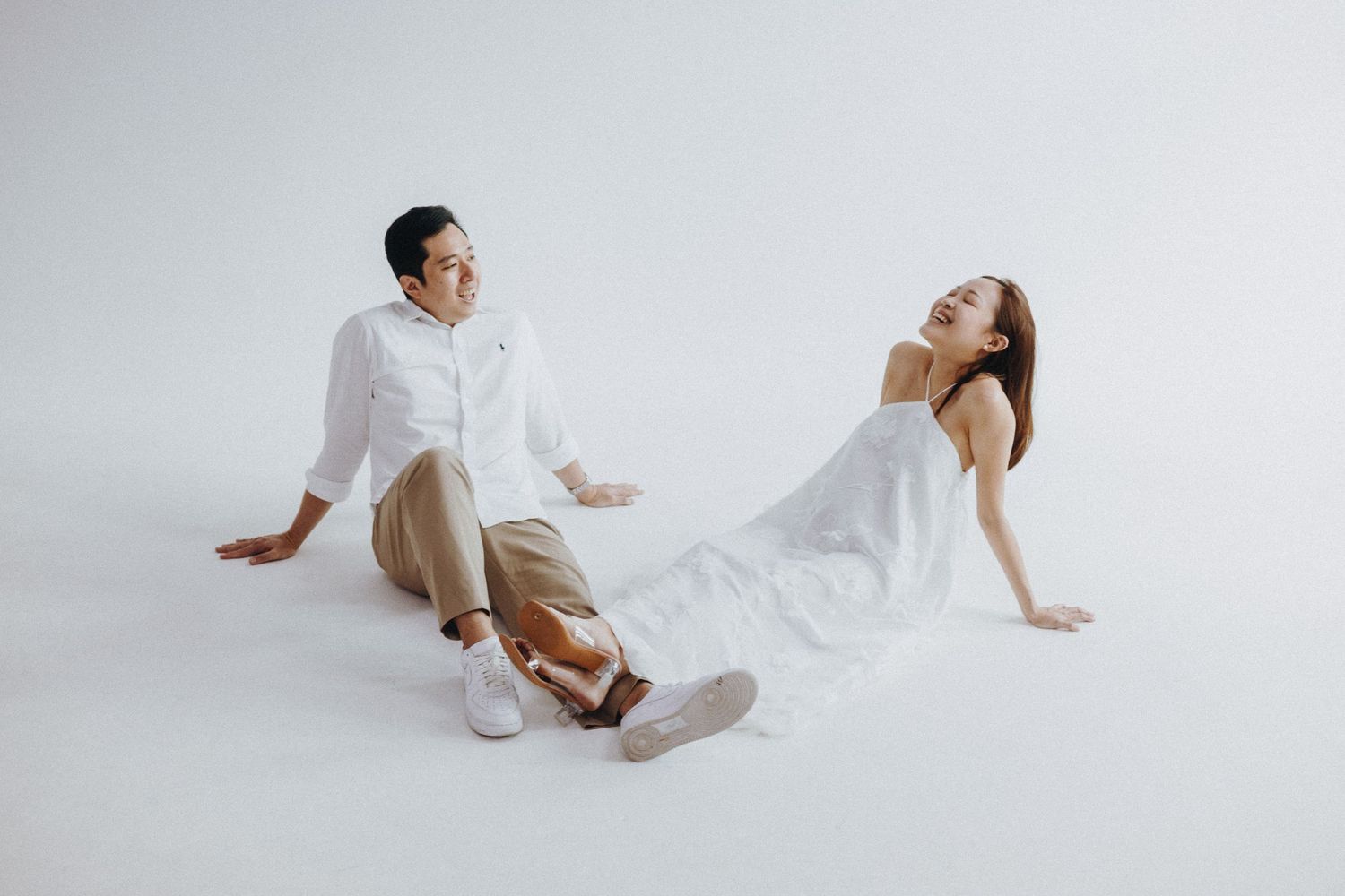 Two people in white and beige attire pose gracefully against a white backdrop during a minimalist photoshoot.