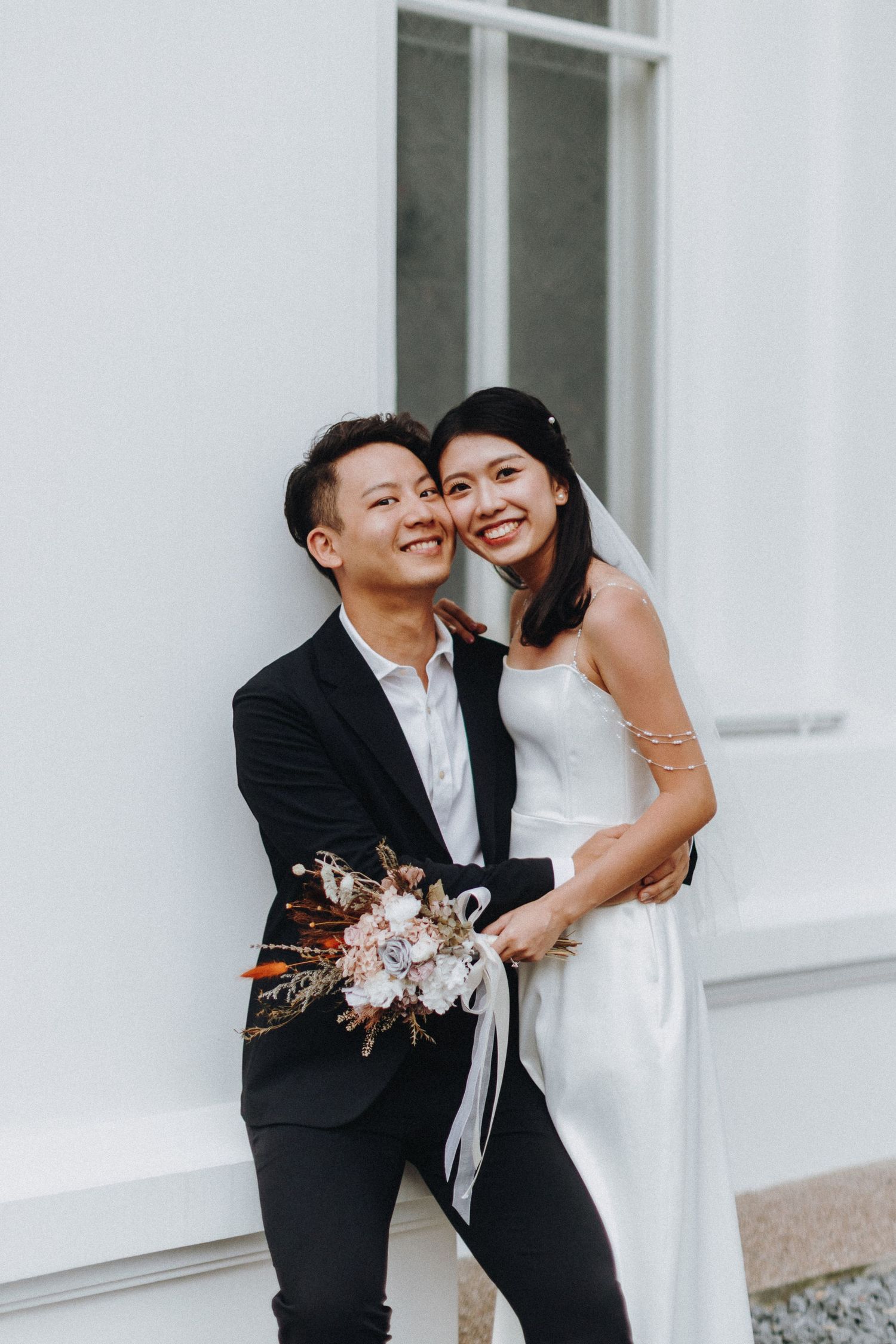 A joyful wedding couple shares a candid moment against a white wall while holding a delicate floral bouquet.