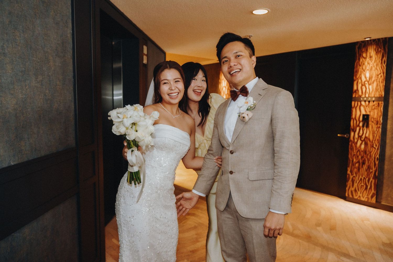 Elegant couple in wedding attire posing with white bouquet in hotel hallway.