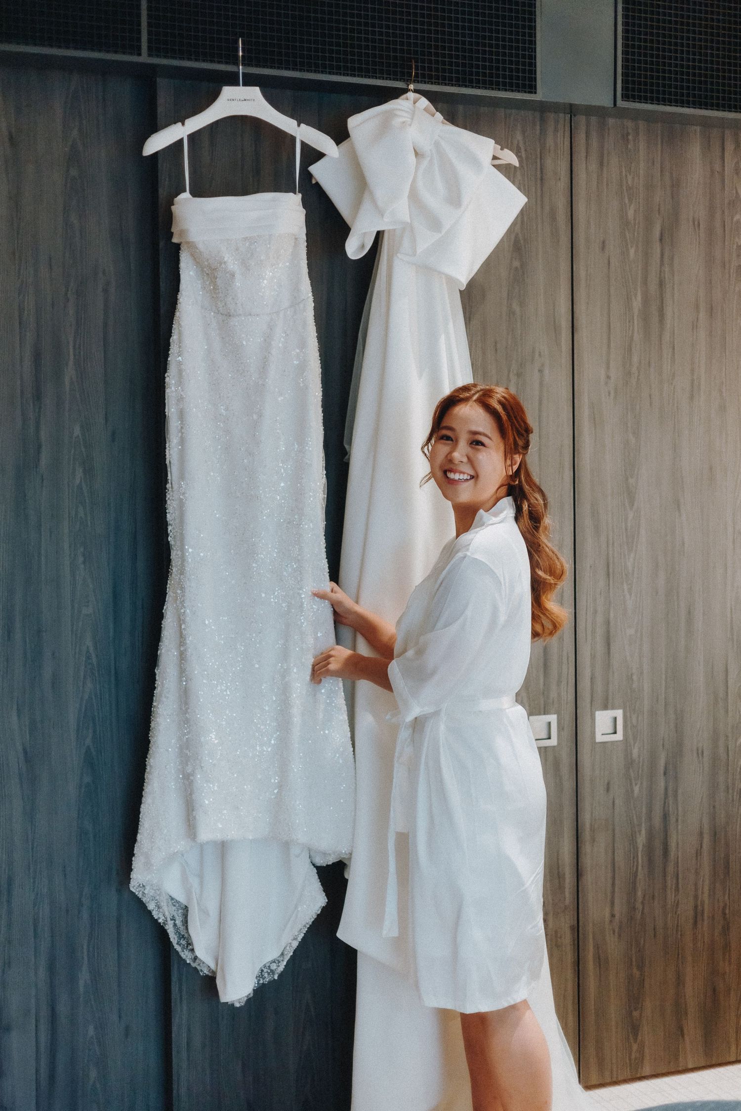 A person in a white robe admires two wedding dresses hanging against a wooden wall.