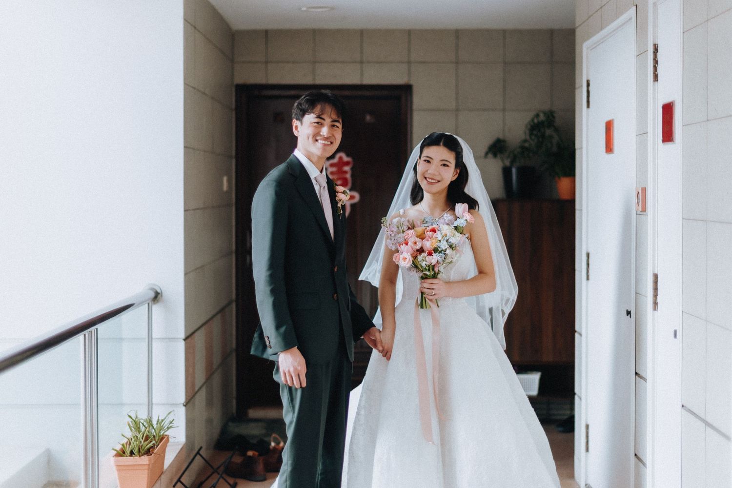 A smiling groom in a dark suit and bride in a white wedding dress holding a pink bouquet pose together in a hallway.