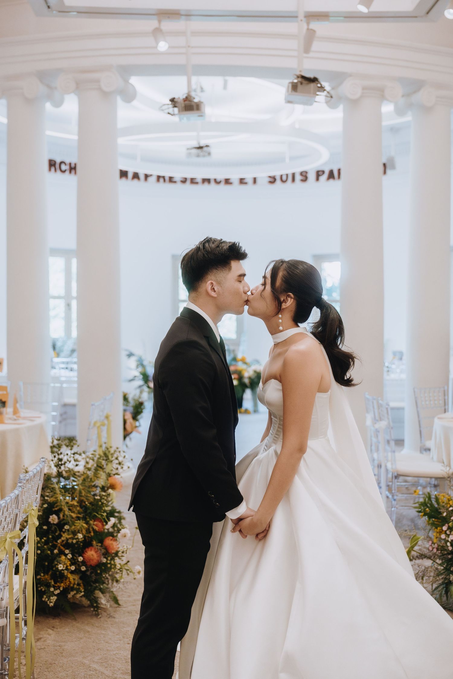 Bride and groom share a kiss at the altar surrounded by floral arrangements and white columns.