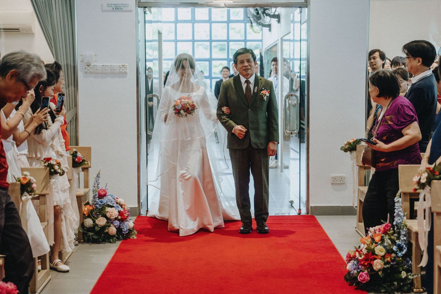 Bride in white gown walks down red carpet aisle with escort at elegant church wedding ceremony.
