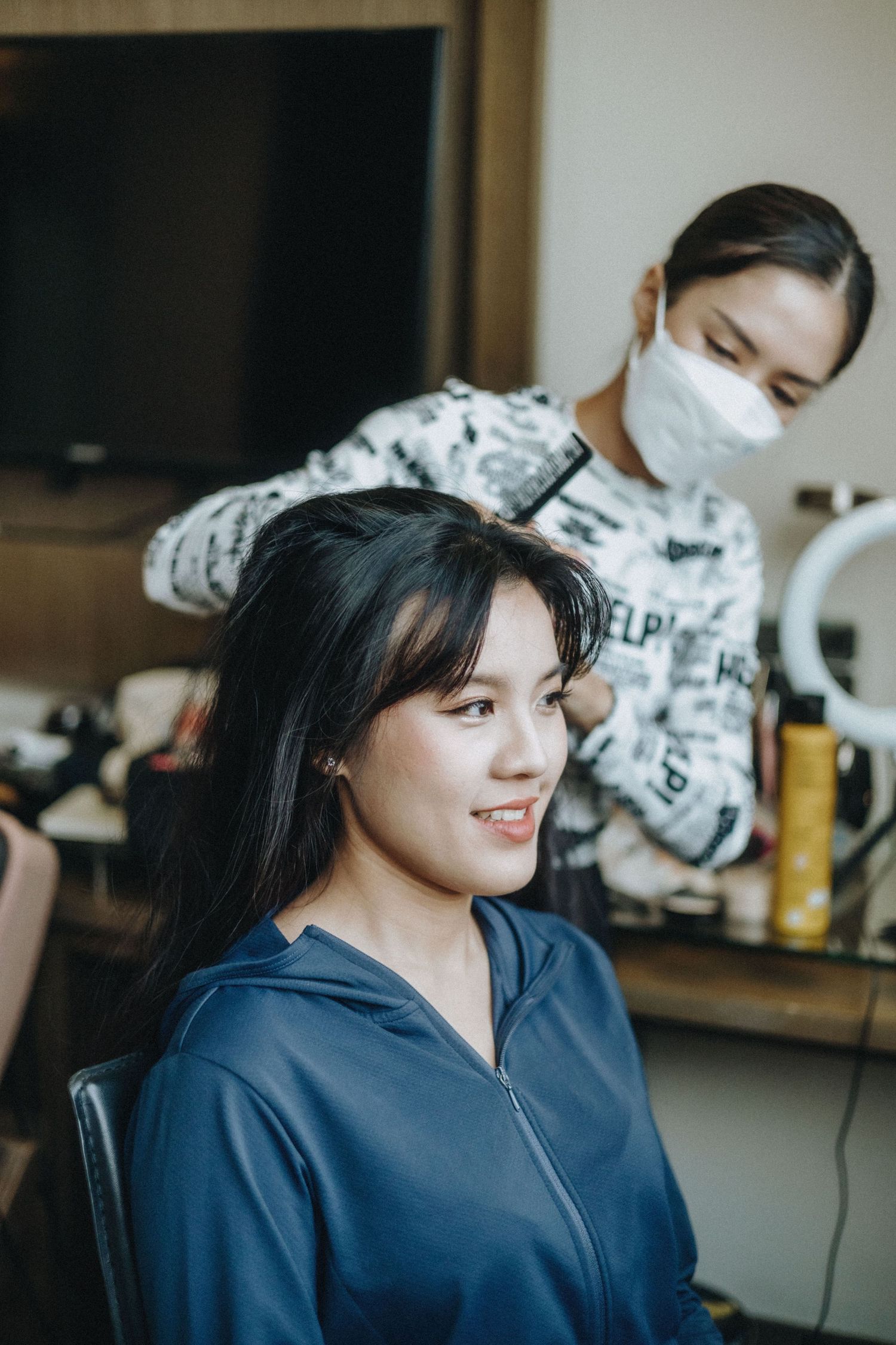 A hairstylist wearing a mask styles the dark hair of a smiling young woman in a blue jacket.