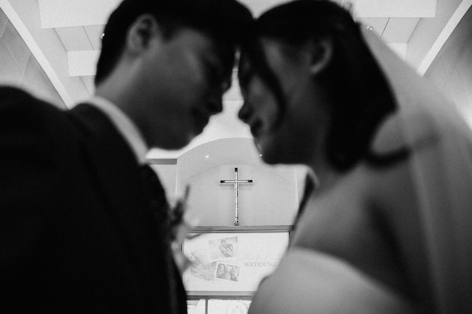 Bride and groom touch foreheads intimately in a black and white church wedding photo with a cross in the background.