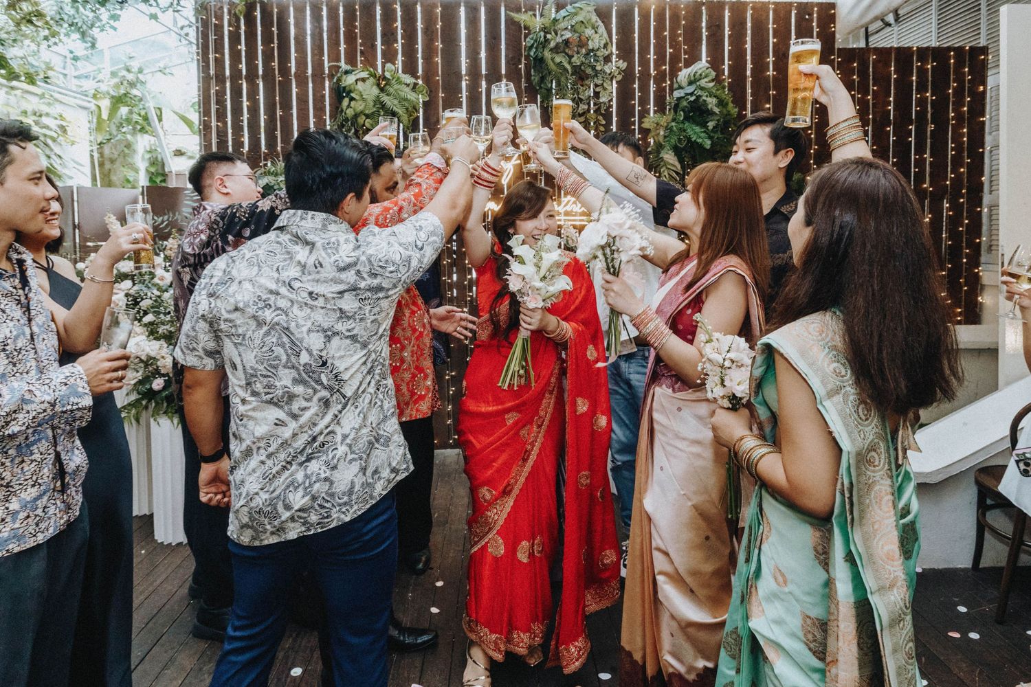 Wedding guests raising glasses in a celebratory toast around the bride in a red saree outdoors.